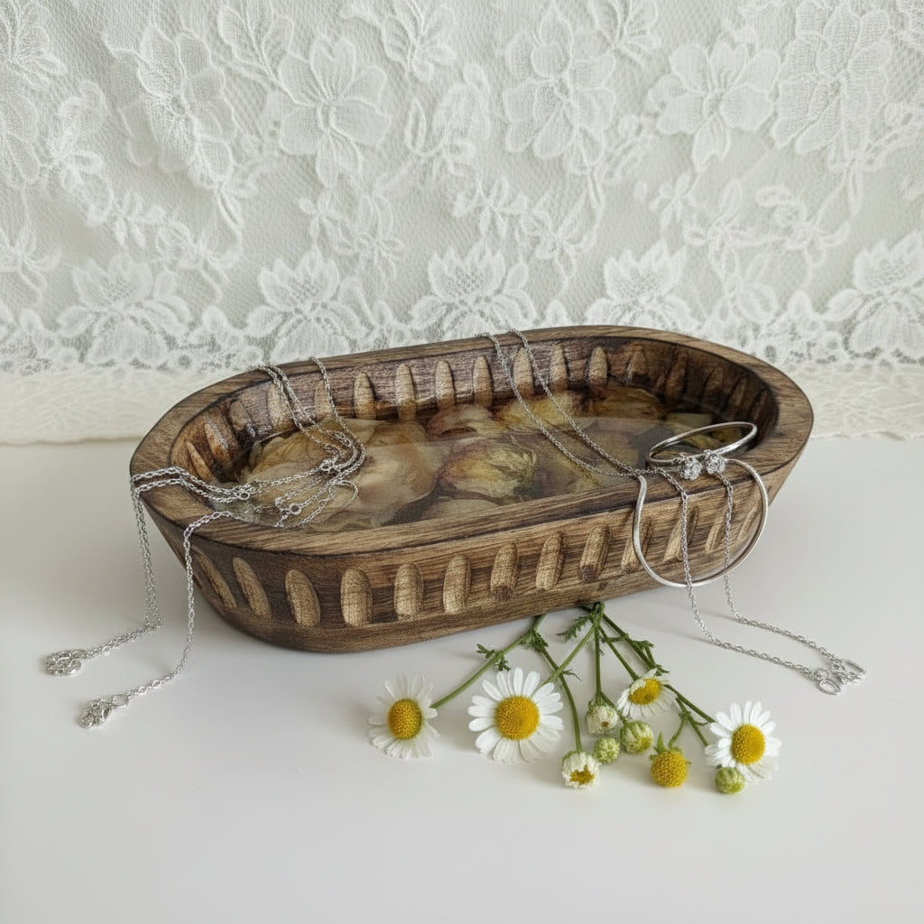 An oval wooden tray with a woven basket-like edge, filled with dried flowers and botanical pieces, placed on a white surface in front of a white shelf. White daisies laying up in front. 