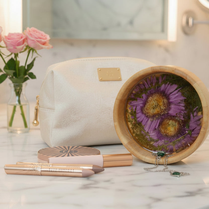 A cream-colored faux-leather cosmetic pouch with a gold-tone logo plate sits on a marble countertop next to a round wooden bowl containing dried purple flowers, with two rose-gold lipsticks and a compact mirror in front.