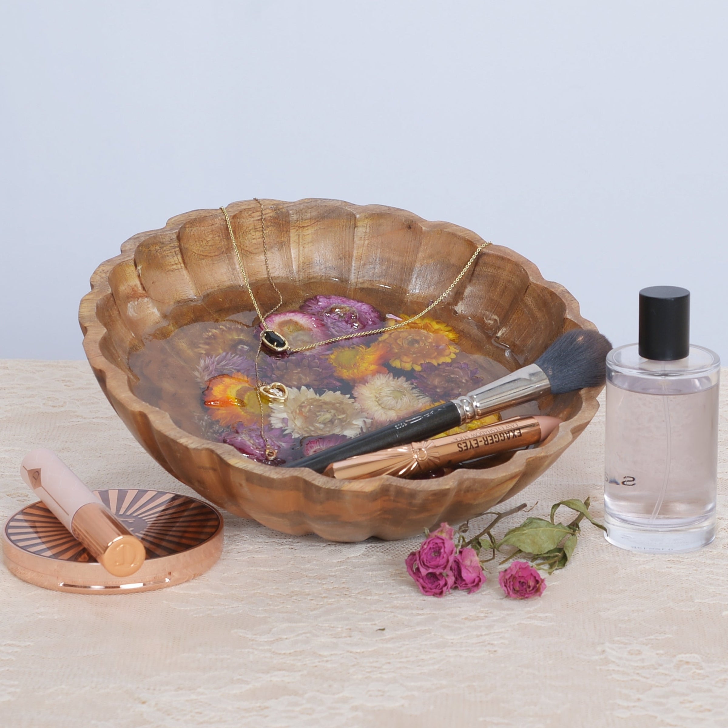 A scalloped wooden bowl filled with clear resin and dried flowers, and makeup brushes, styled on a light beige textured surface next to a clear bottle with a black cap and a pink makeup sponge.
