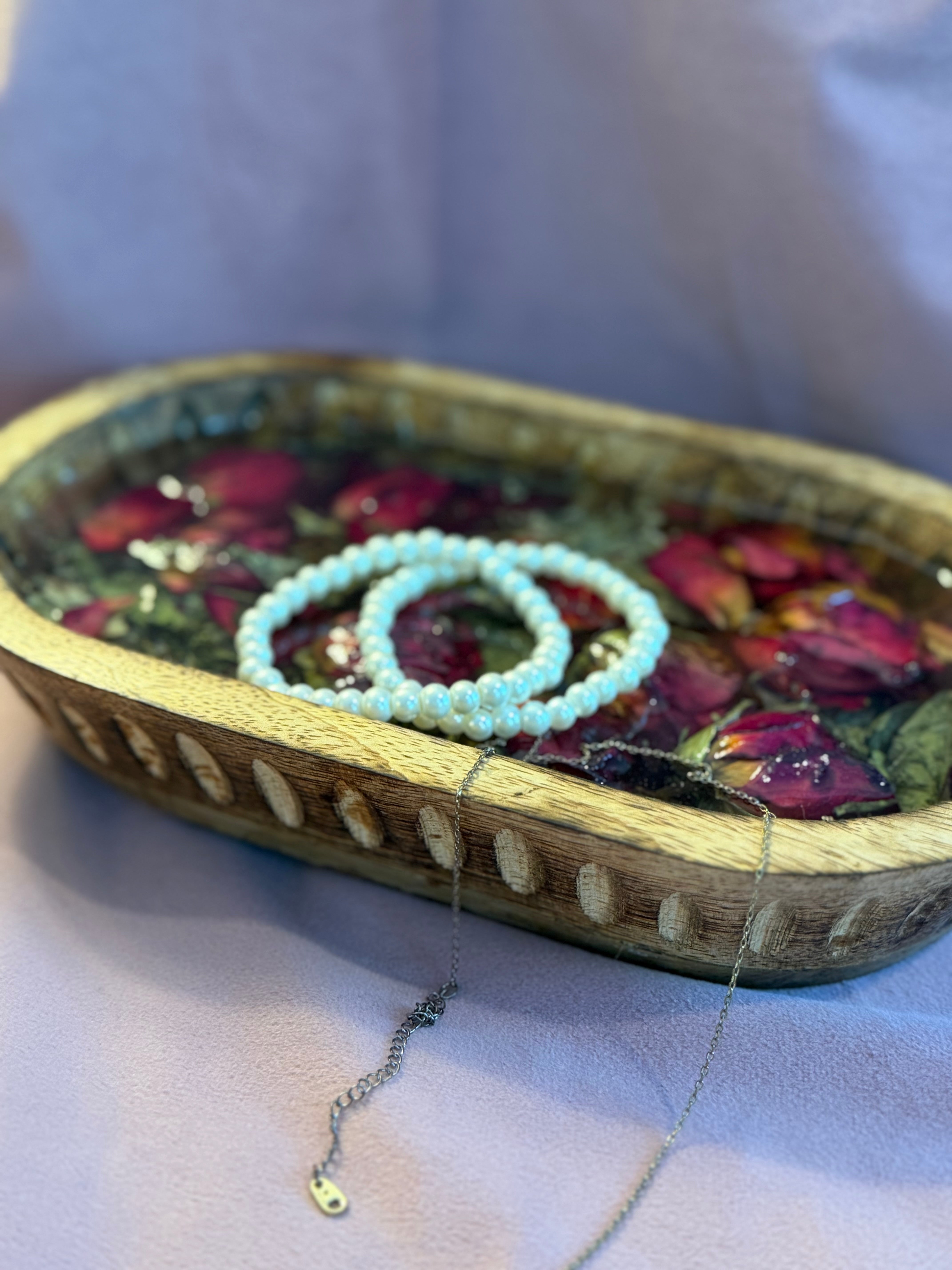 Pearl bracelets and a delicate chain necklace draped over a carved wooden dough bowl filled with red rosebuds in resin