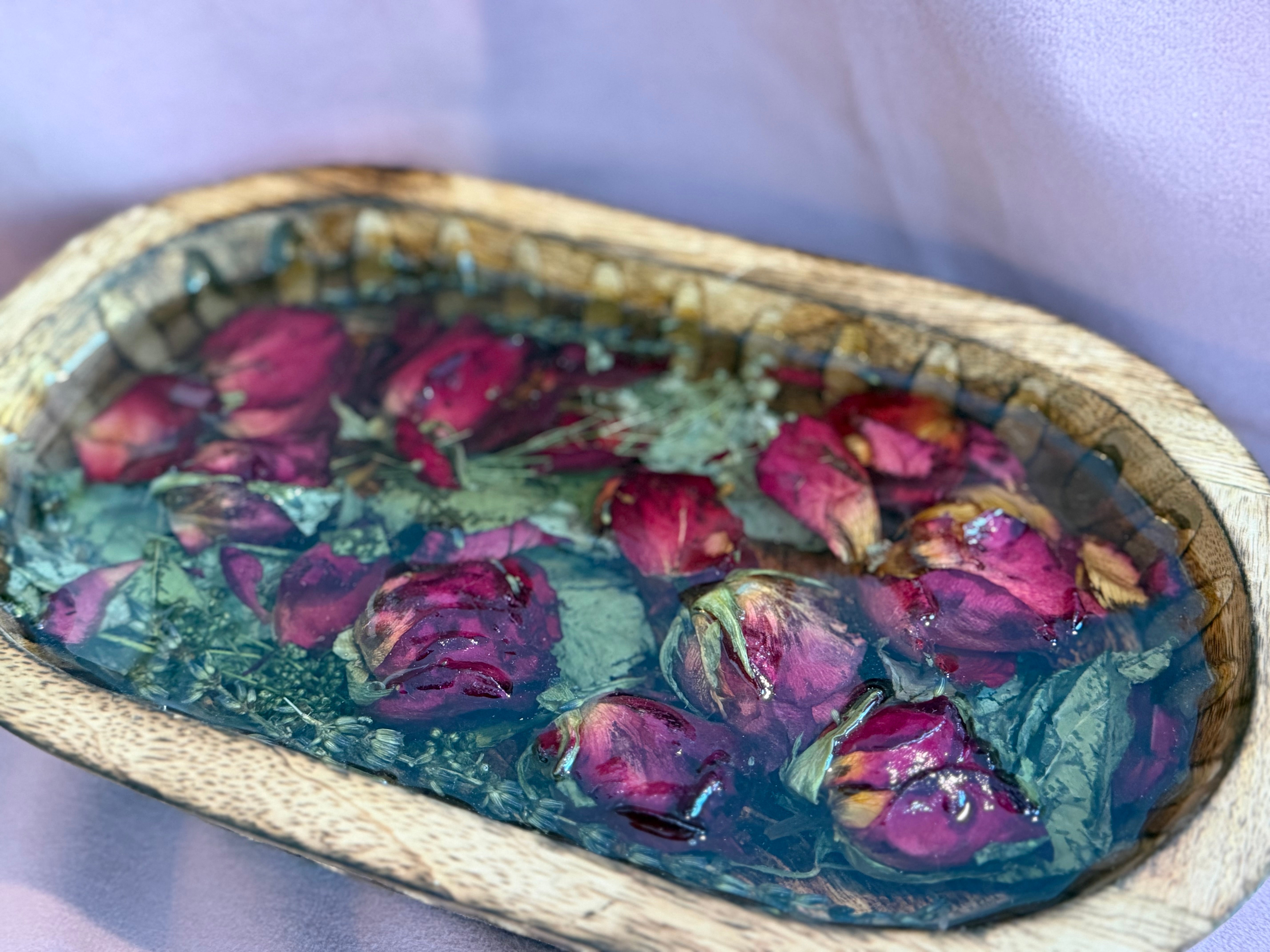 Close-up of dried red rosebuds, green leaves, and herbs set in glass-clear resin inside a carved wooden dough bowl