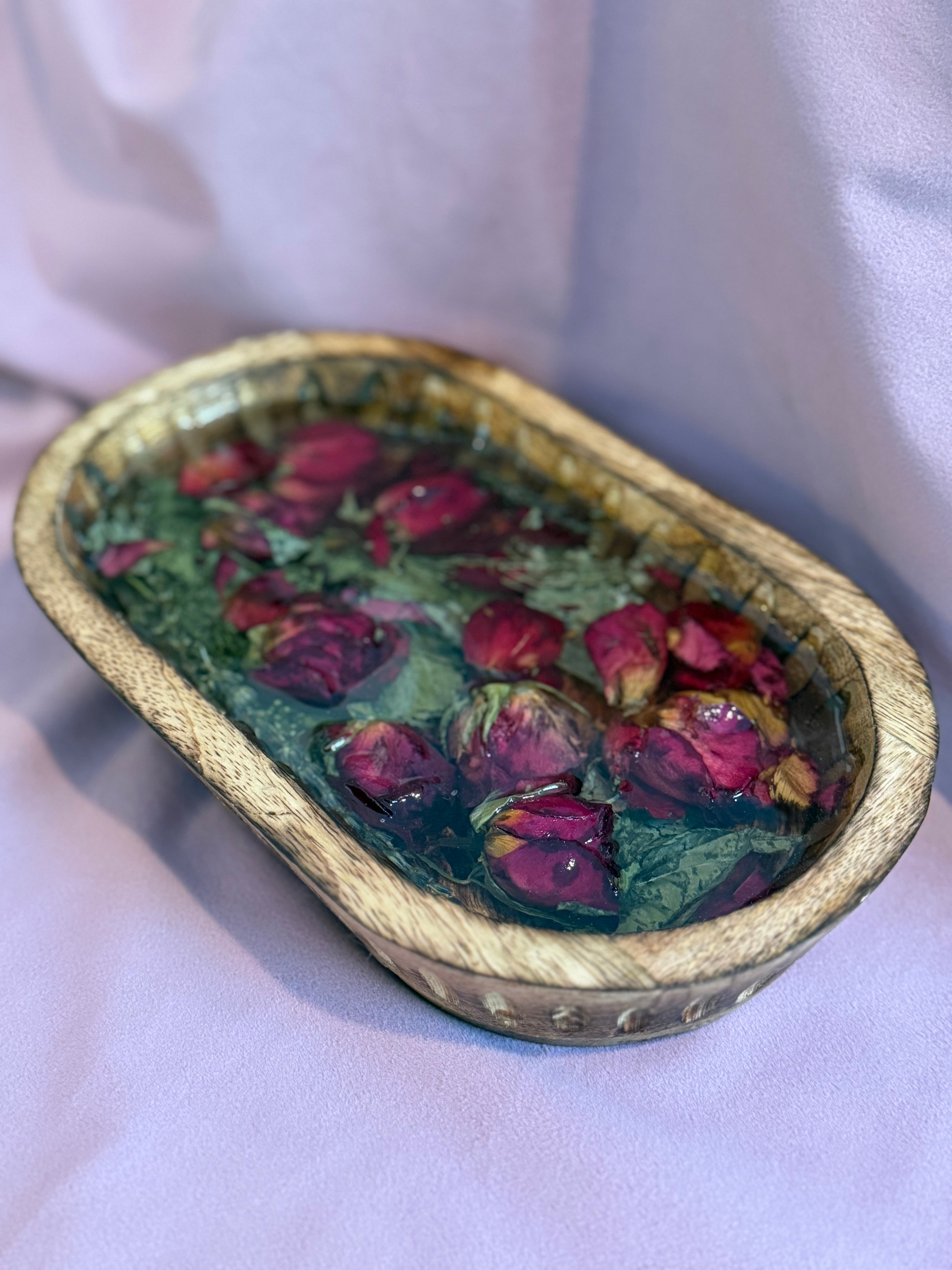 Hand-carved oval wooden dough bowl filled with deep red rosebuds, green leaves, and baby's breath sealed in clear resin, on lavender fabric