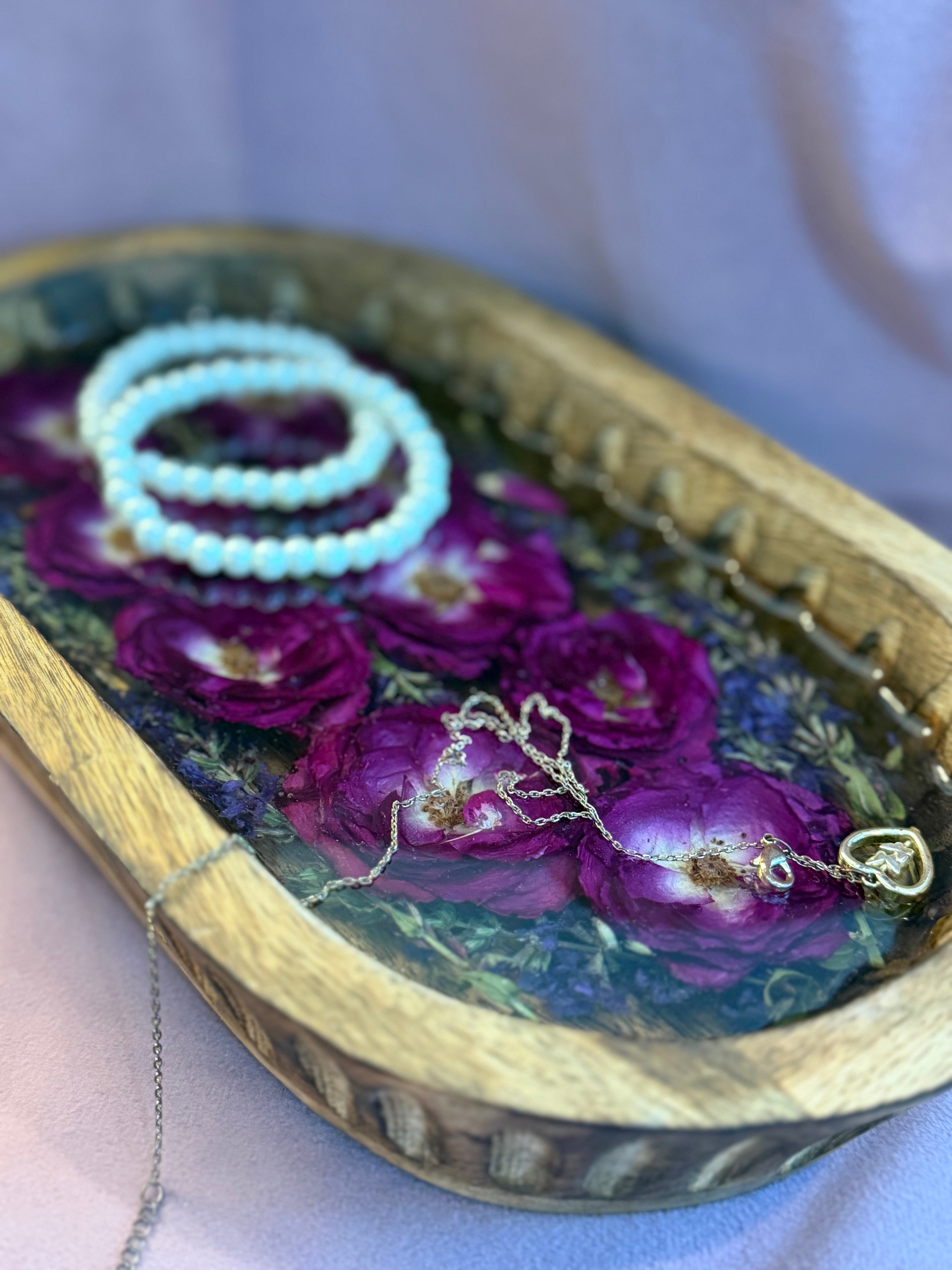 Close-up of magenta roses and purple botanicals in resin inside a carved dough bowl, styled with pearl bracelets and a gold heart necklace