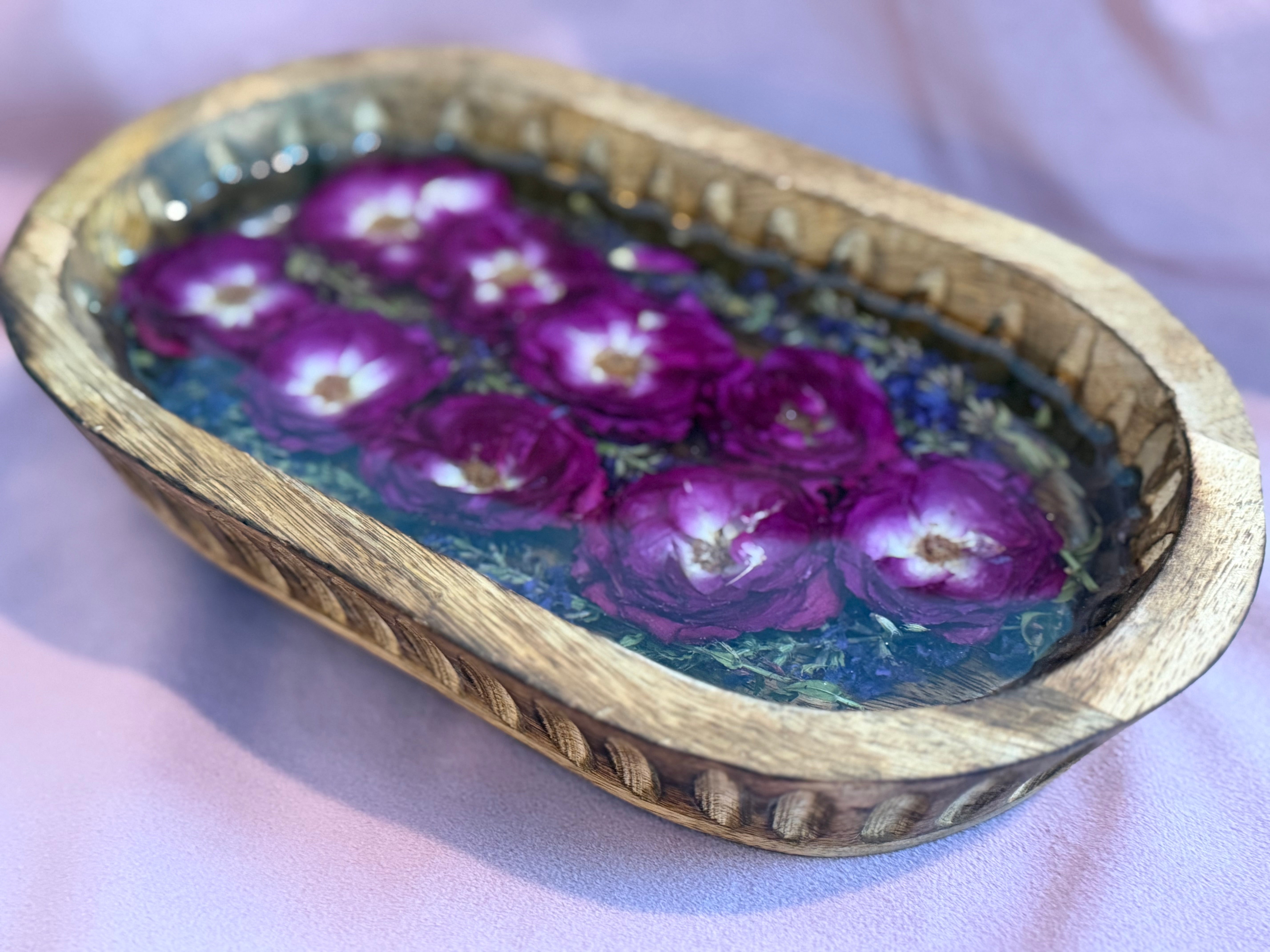 Hand-carved oval wooden dough bowl filled with magenta roses with white centers, purple blooms, and greenery sealed in clear resin, on lavender fabric