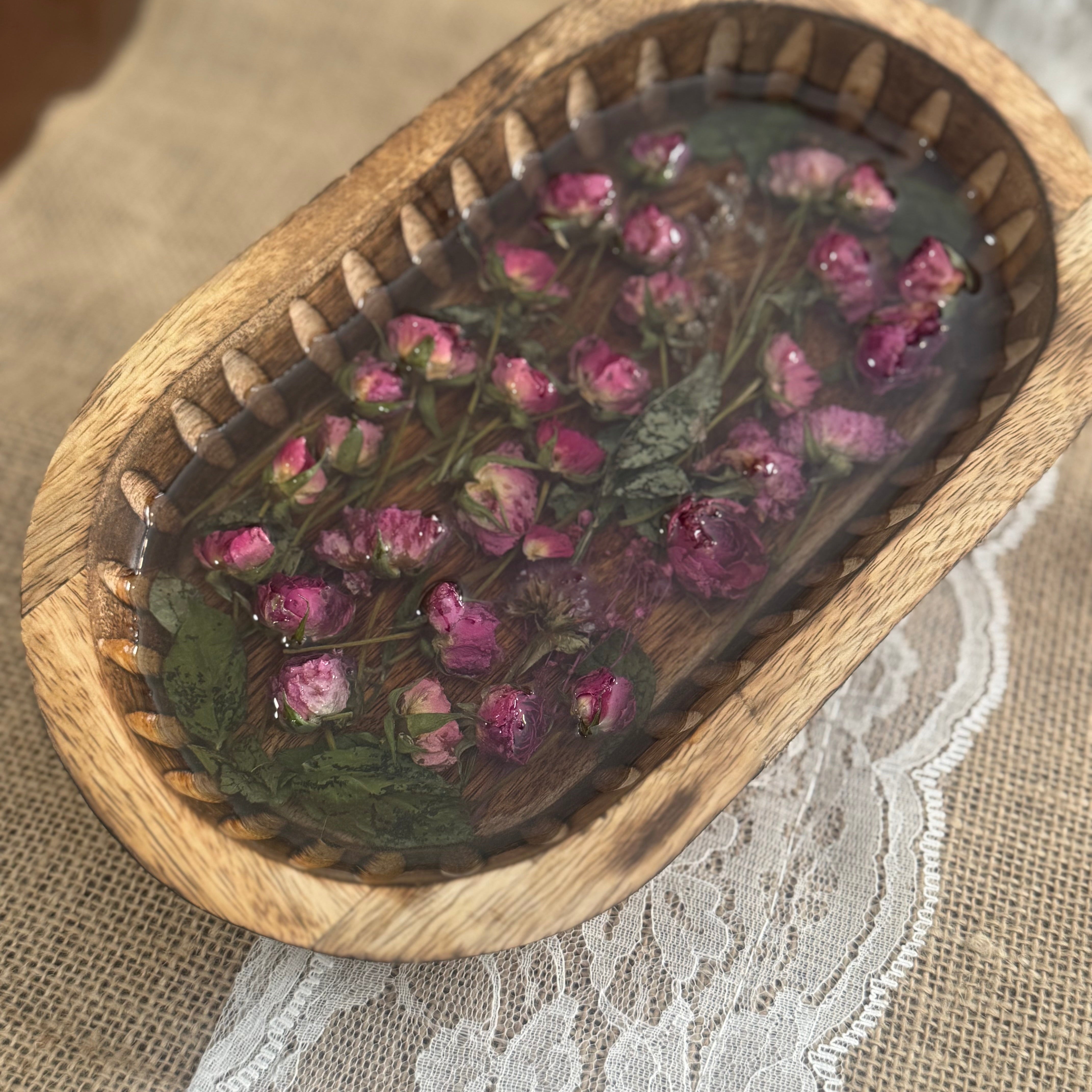 An oval wooden tray filled with dried pink mini roses and green leaves set in clear resin, placed on a beige fabric surface with white lace.