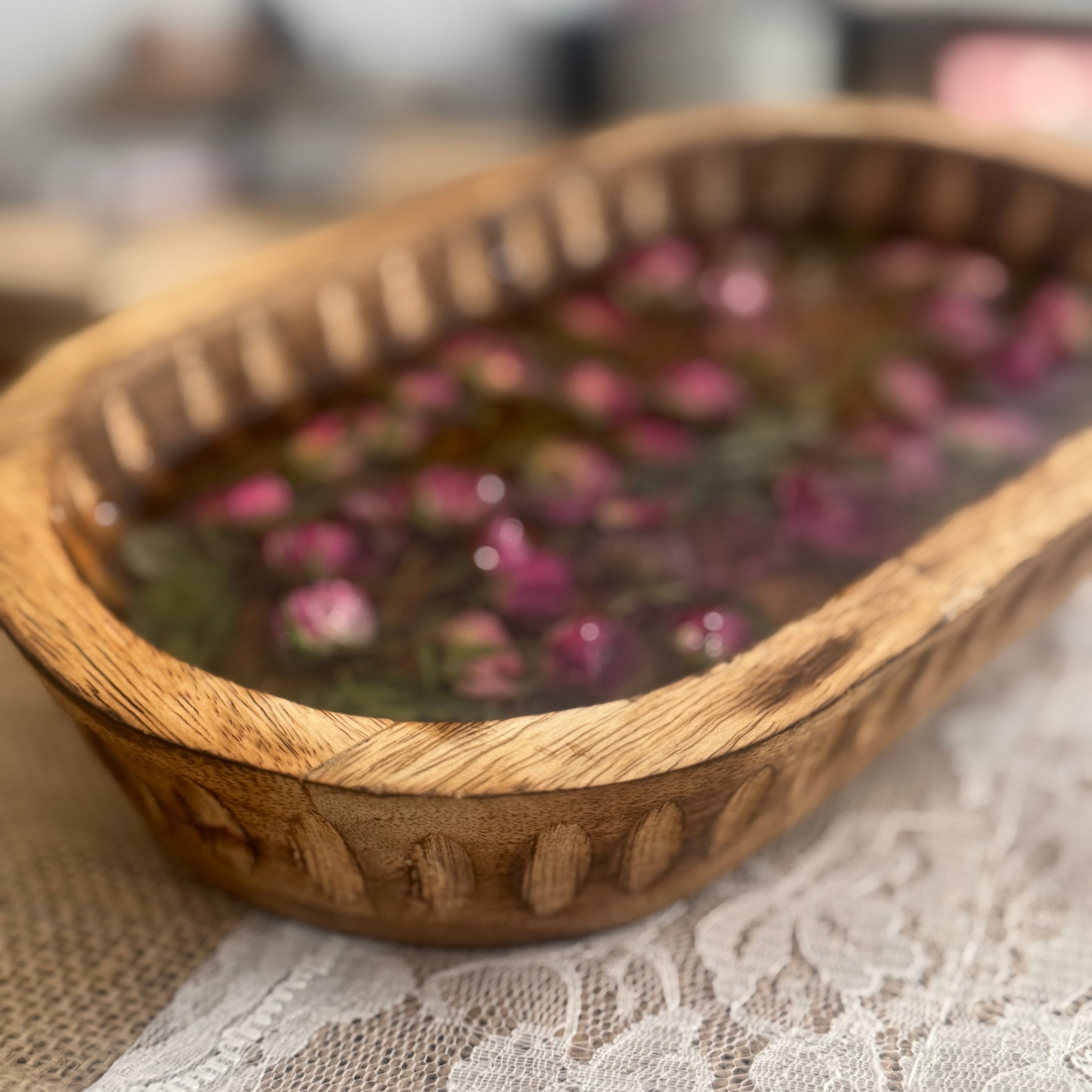 A close-up of a carved wooden bowl filled with dried pink mini rose petals set in dark-toned clear resin, resting on white lace cloth.