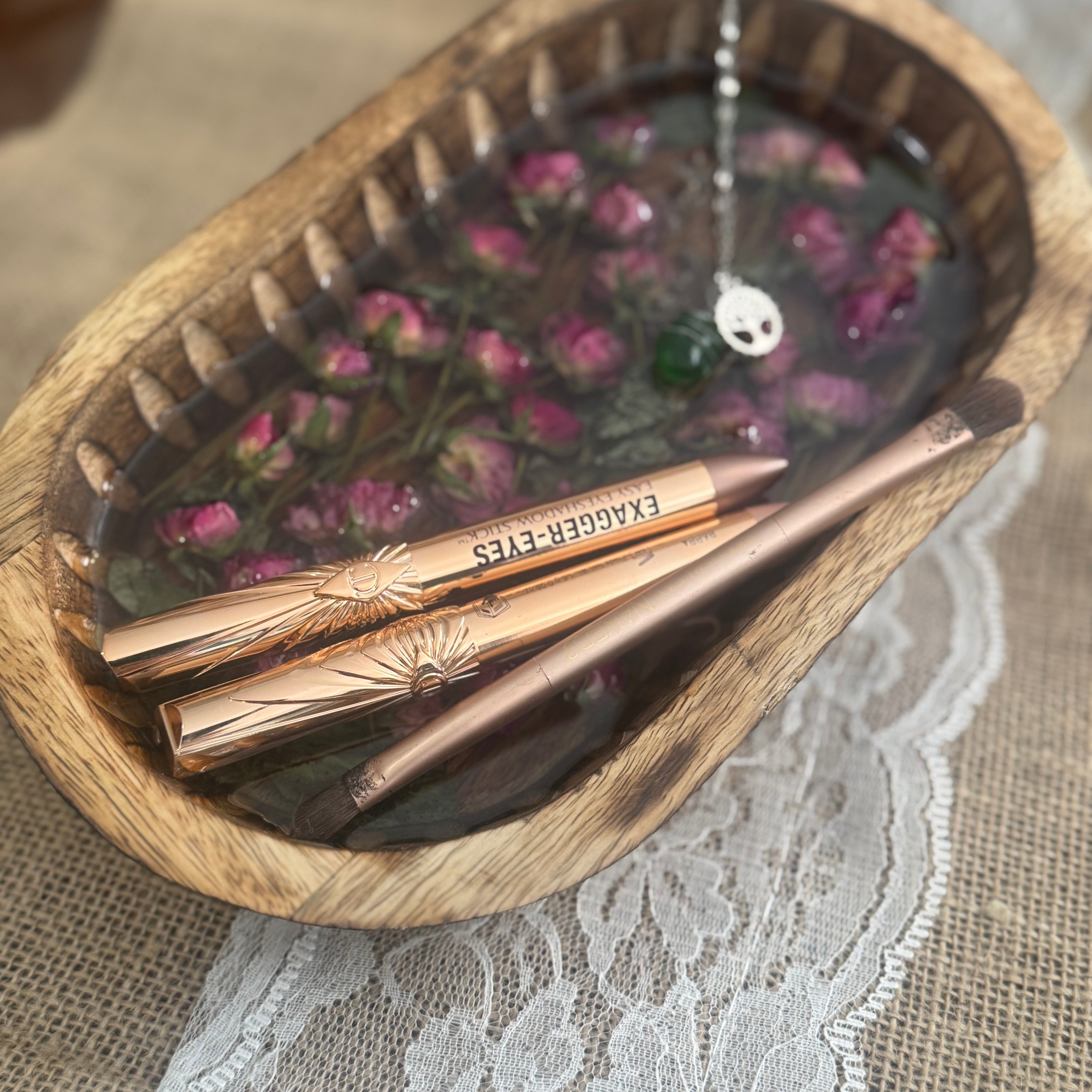 Rose golden eyeshadow pencils resting in a decorative wooden bowl filled with dried pink mini roses, set on a lace and burlap surface.