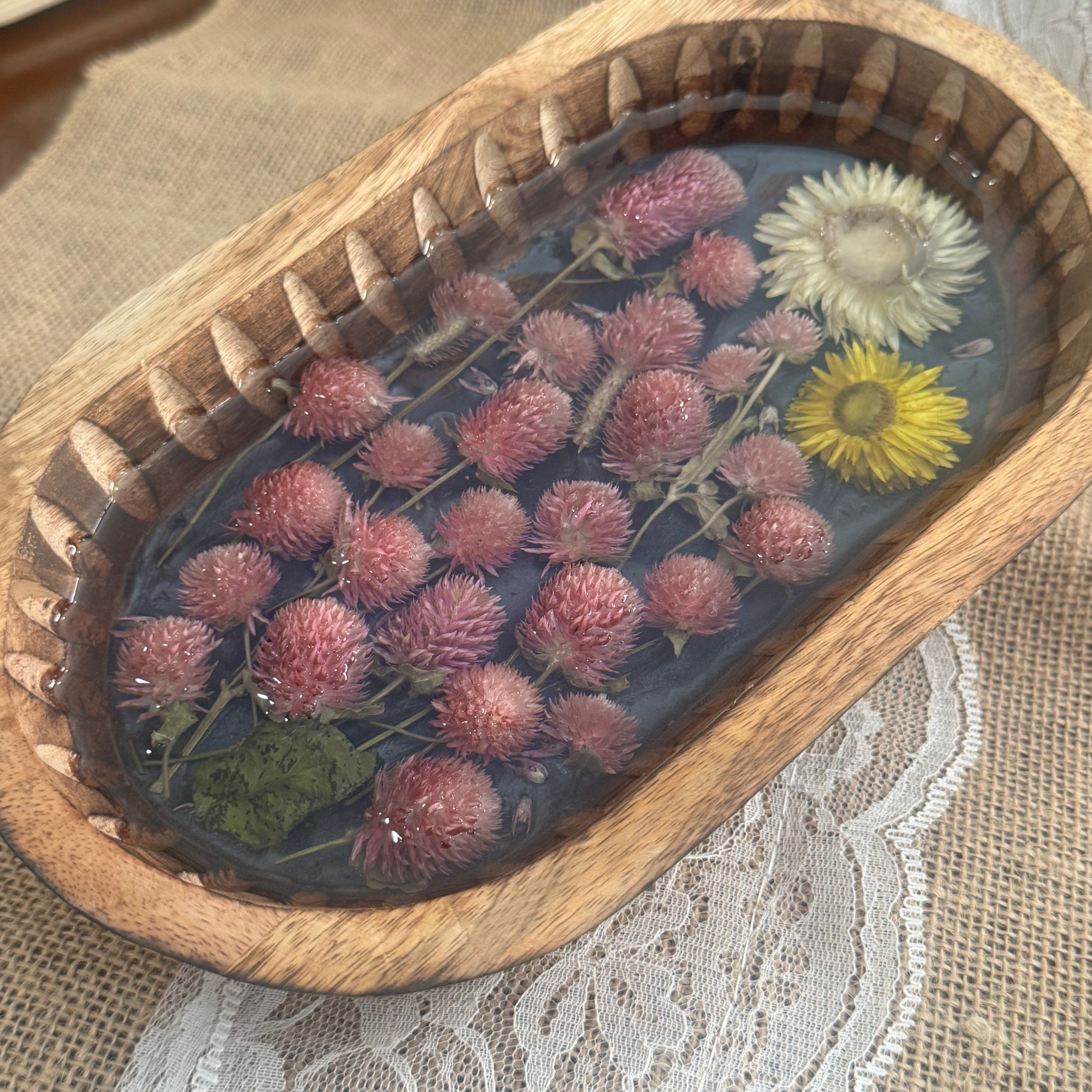 An oval wooden tray filled with preserved dried flowers under clear resin, placed on a beige fabric surface with white lace.