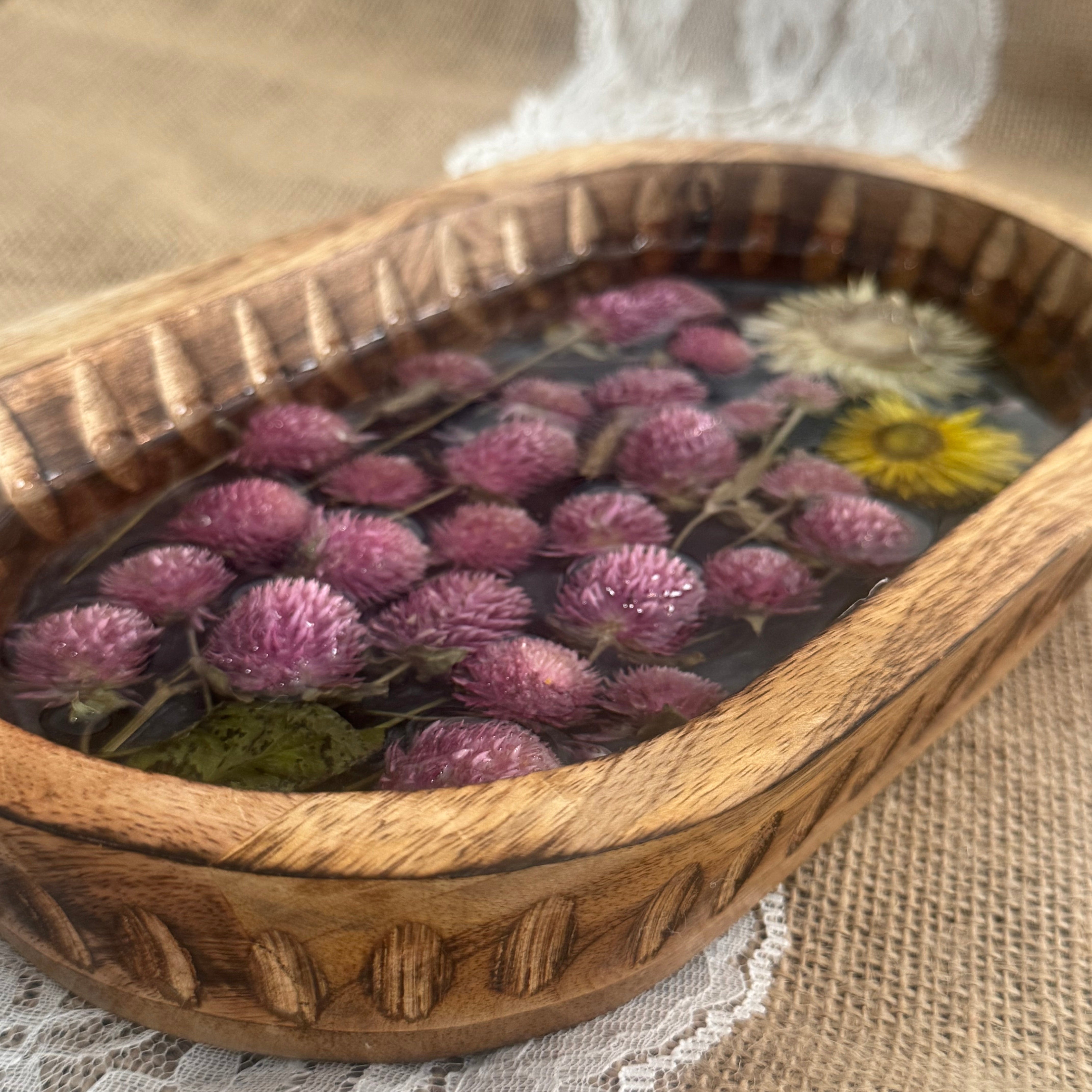 A carved wooden oval bowl with dried pink flowers and a yellow flower, set on a lace cloth over a burlap surface.