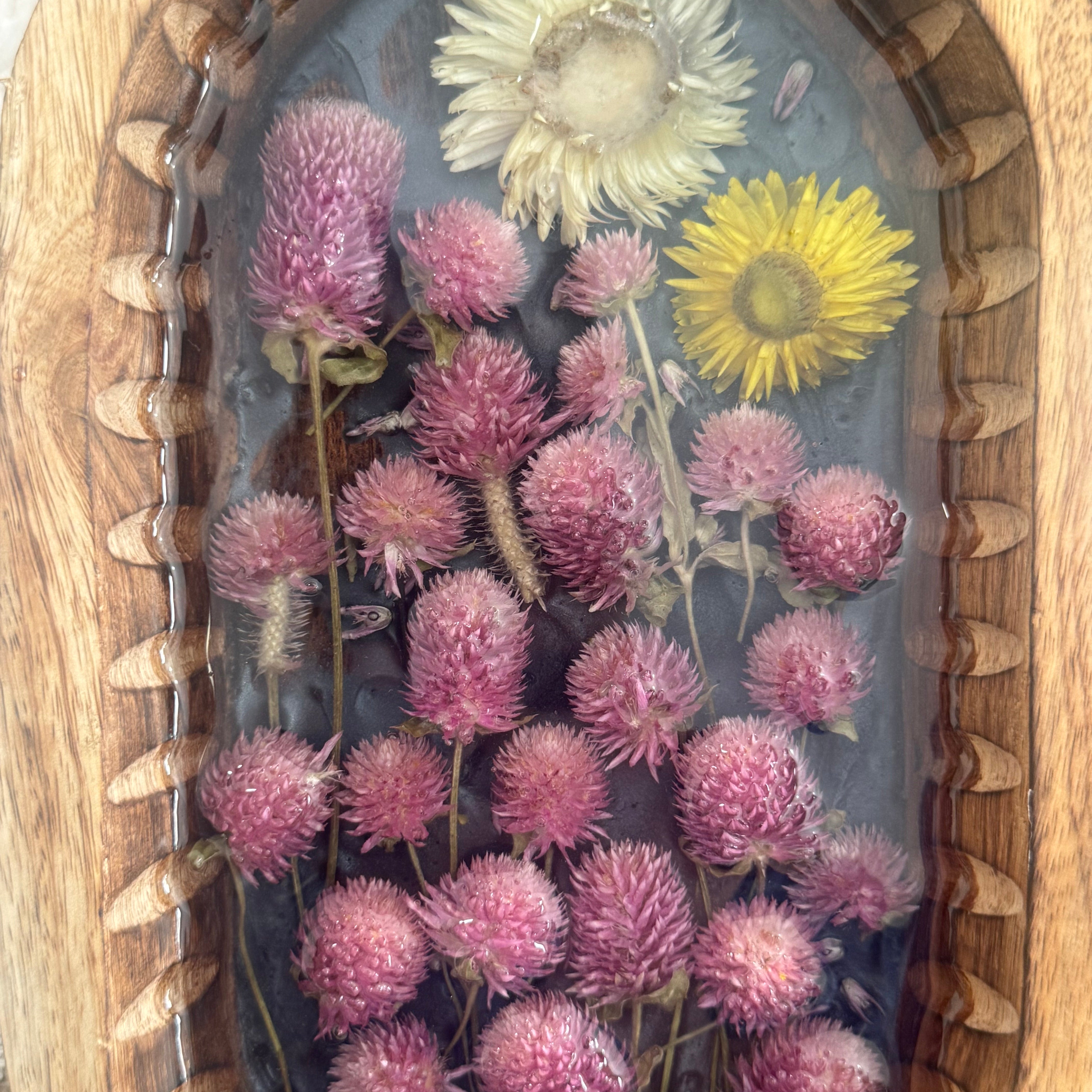 A top-down view of a decorative oval wooden tray containing clear resin, dried pink pom-pom flowers, one yellow flower, and one white flower.