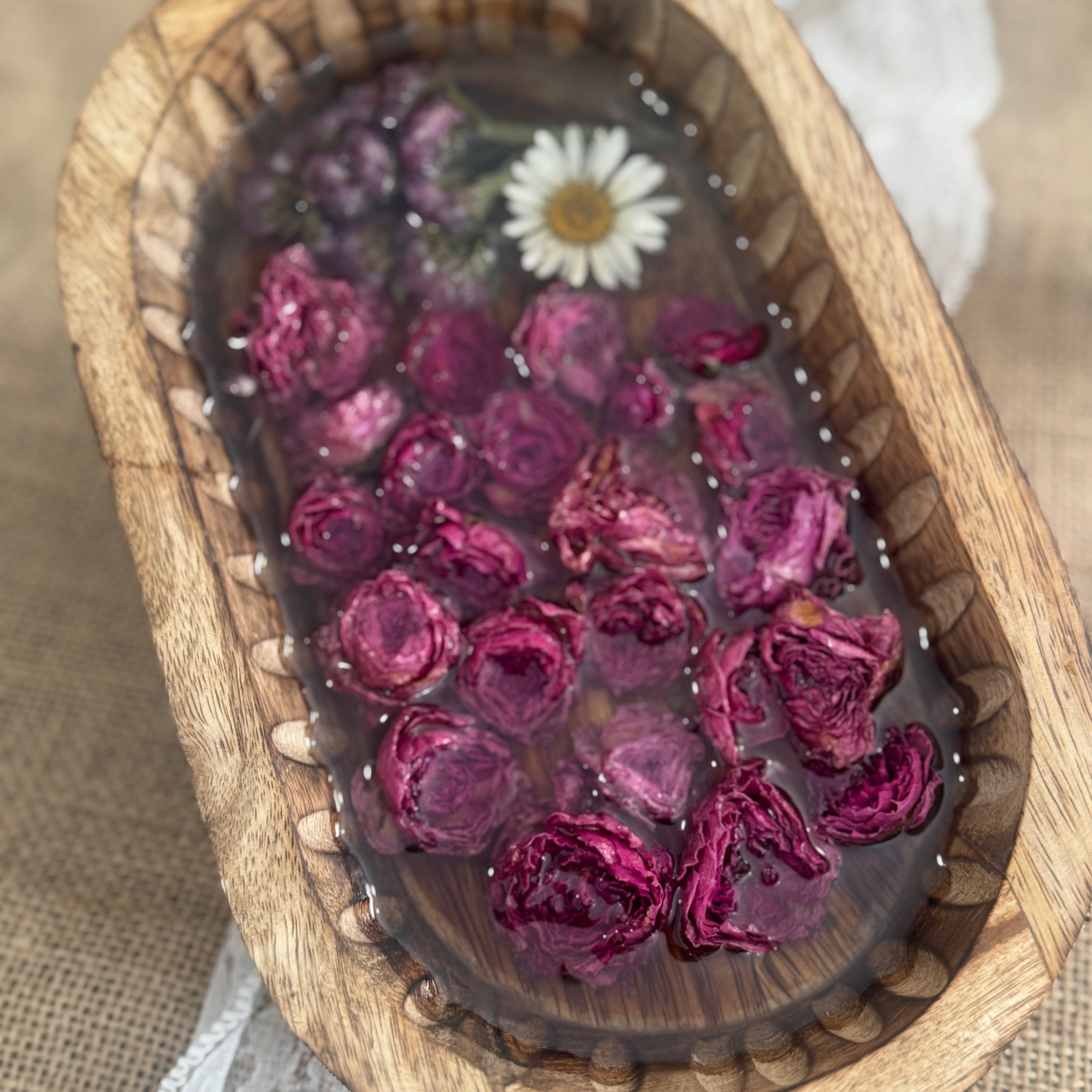 An oval wooden tray filled with a clear resin or gel containing dried pink rose petals and a small white daisy flower, photographed from above on a beige fabric surface with white lace.