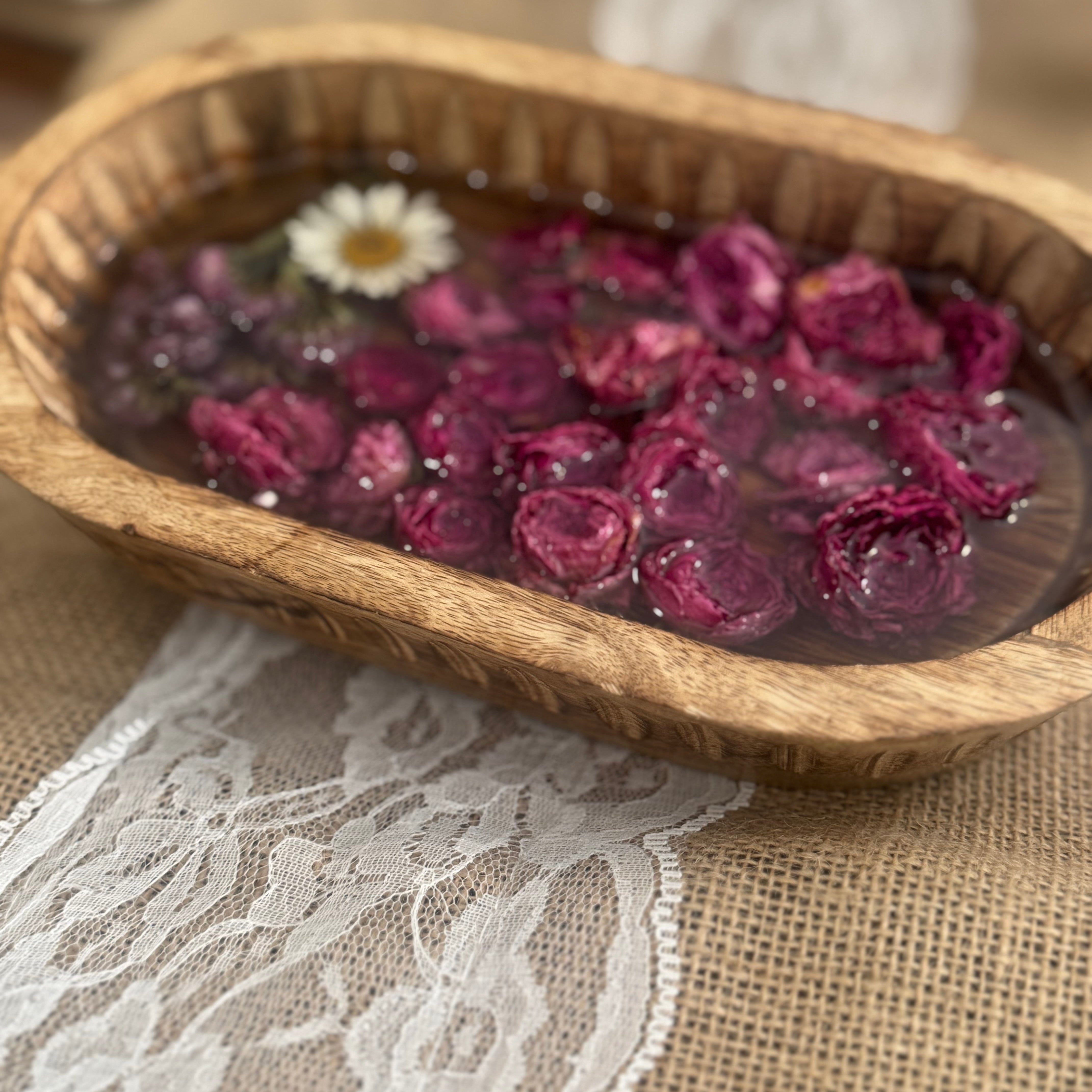 A rustic oval wooden bowl filled with dried pink rose petals and a small white flower set in clear amber-toned resin, on a beige burlap cloth with a white lace runner.