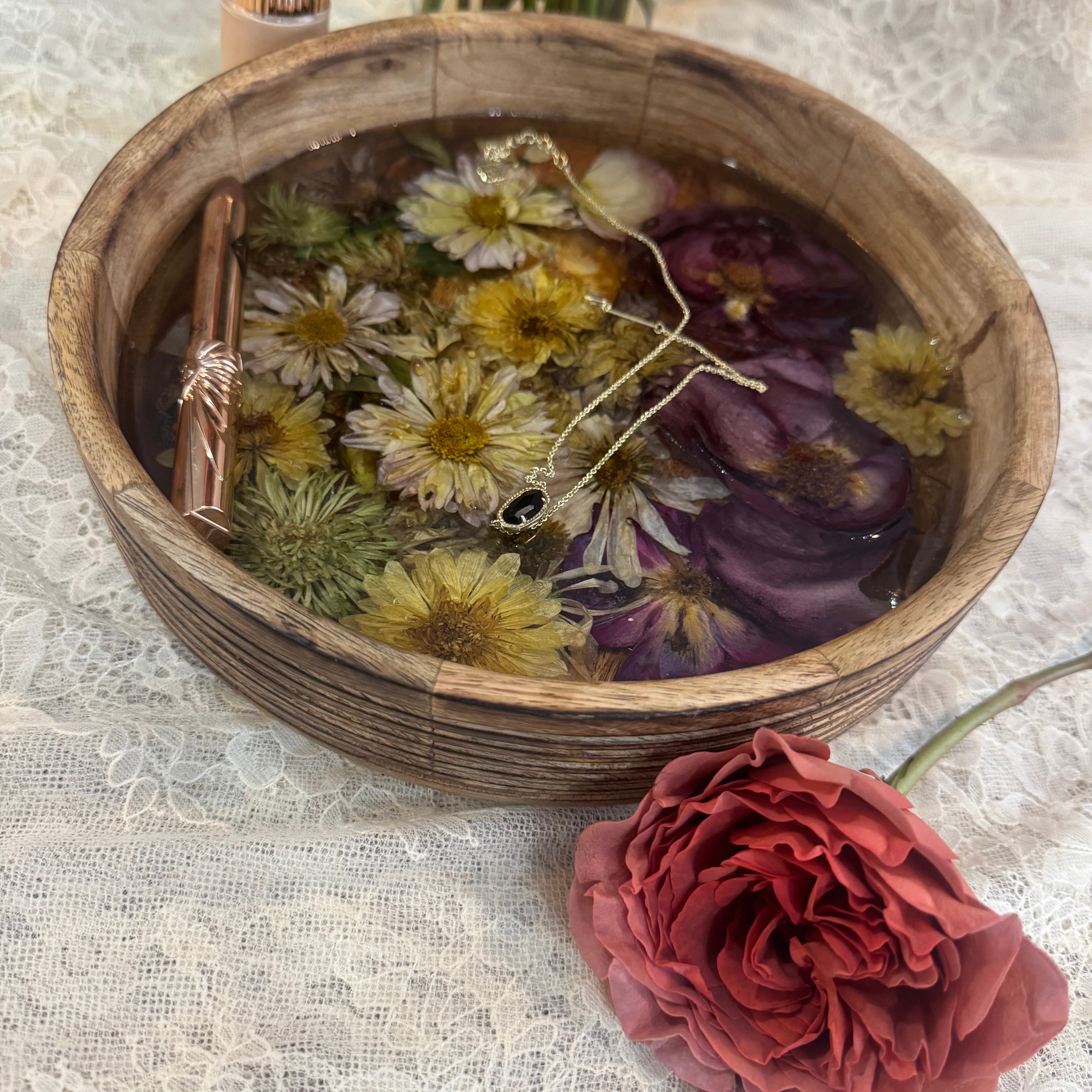 A rustic round wooden bowl filled with dried daisies, wildflowers, and roses set in resin, displayed on a white lace cloth.
