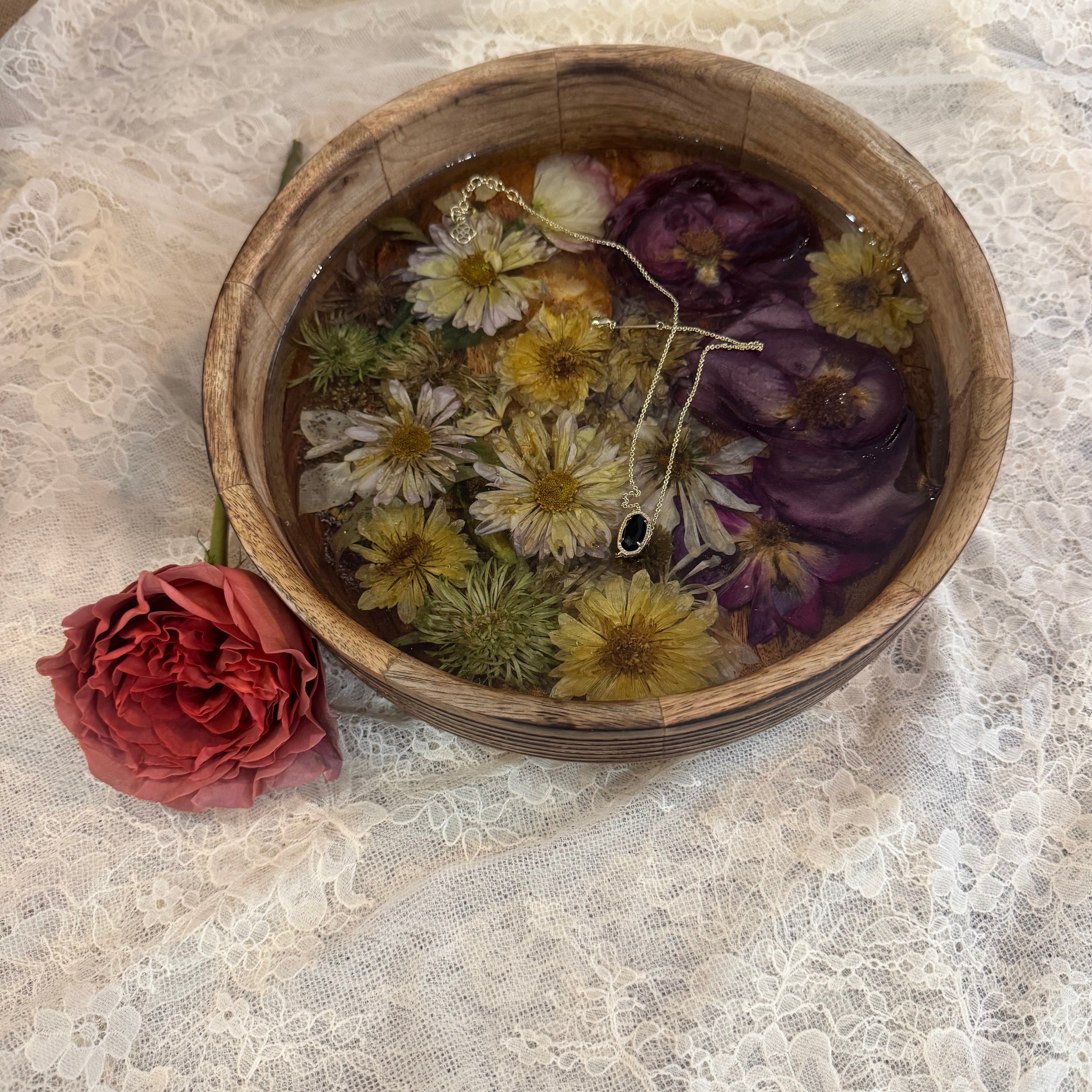 A rustic wooden bowl filled with dried flowers set in clear resin, including white daisies, yellow blossoms, and purple flowers, set on a white lace cloth next to a red rose.