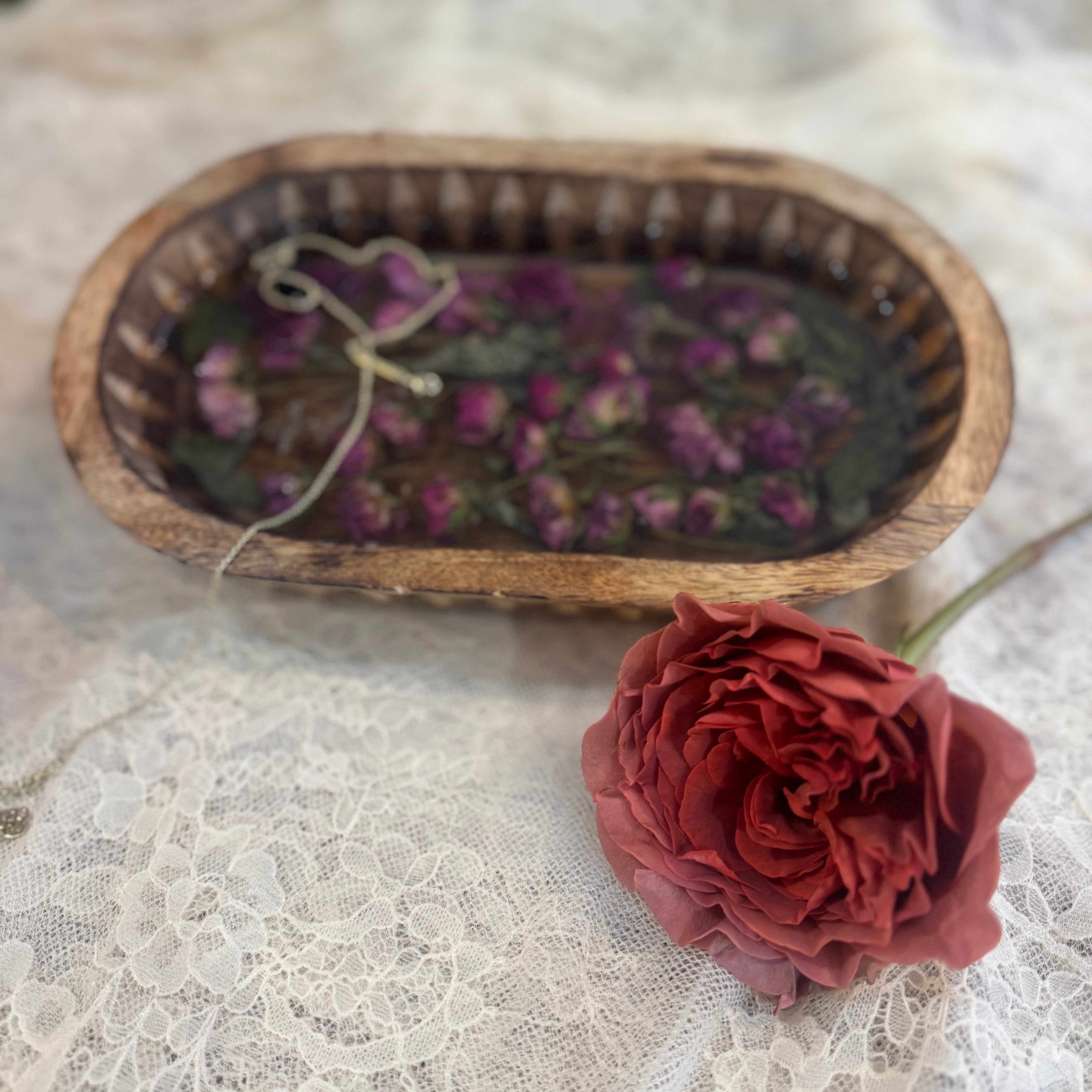A rustic oval wooden tray filled with preserved pink-toned mini roses and green leaves, styled on white lace fabric next to a single red rose.