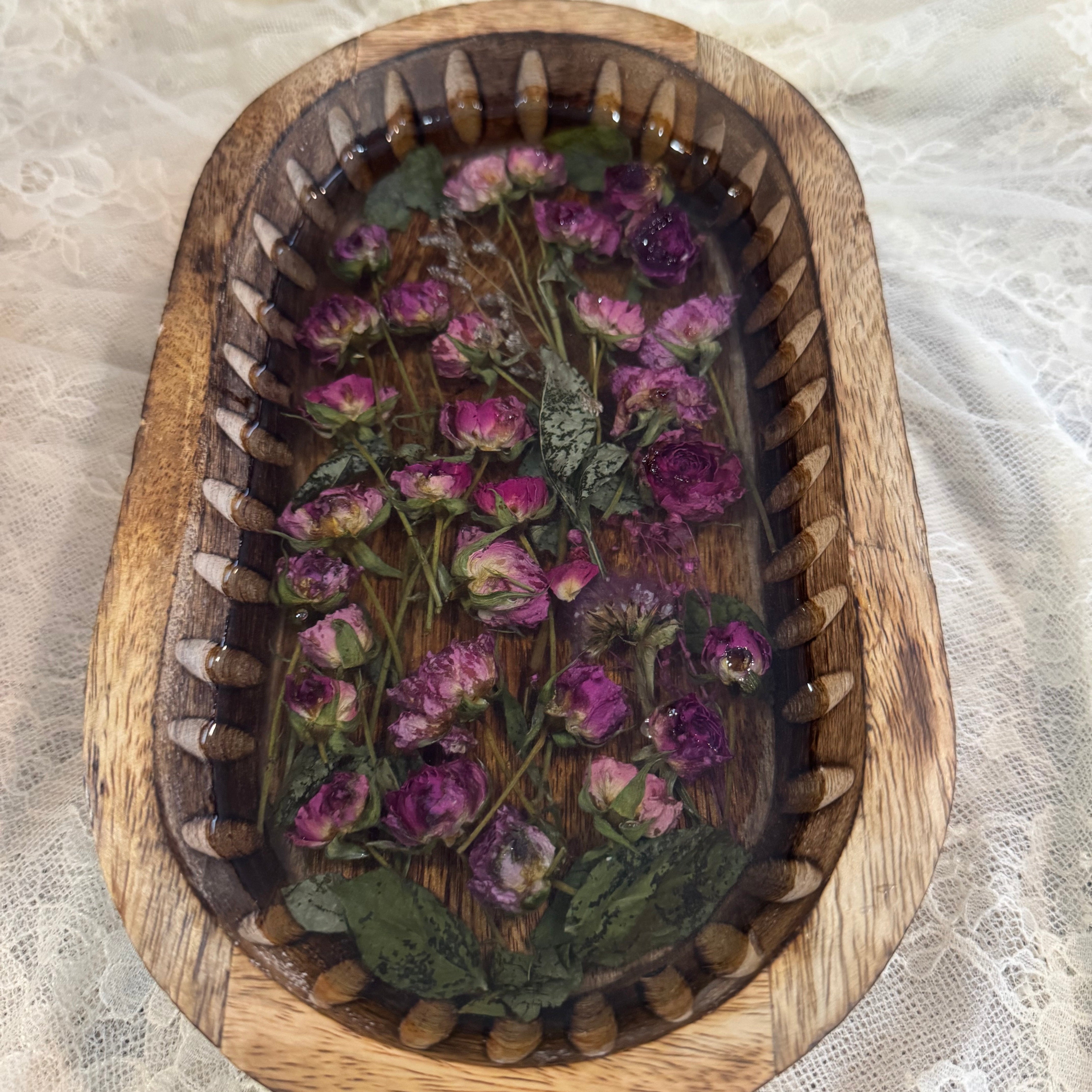 An oval wooden tray with a carved ribbed rim, filled with dried purple flowers and green leaves, set on a white lace fabric background.