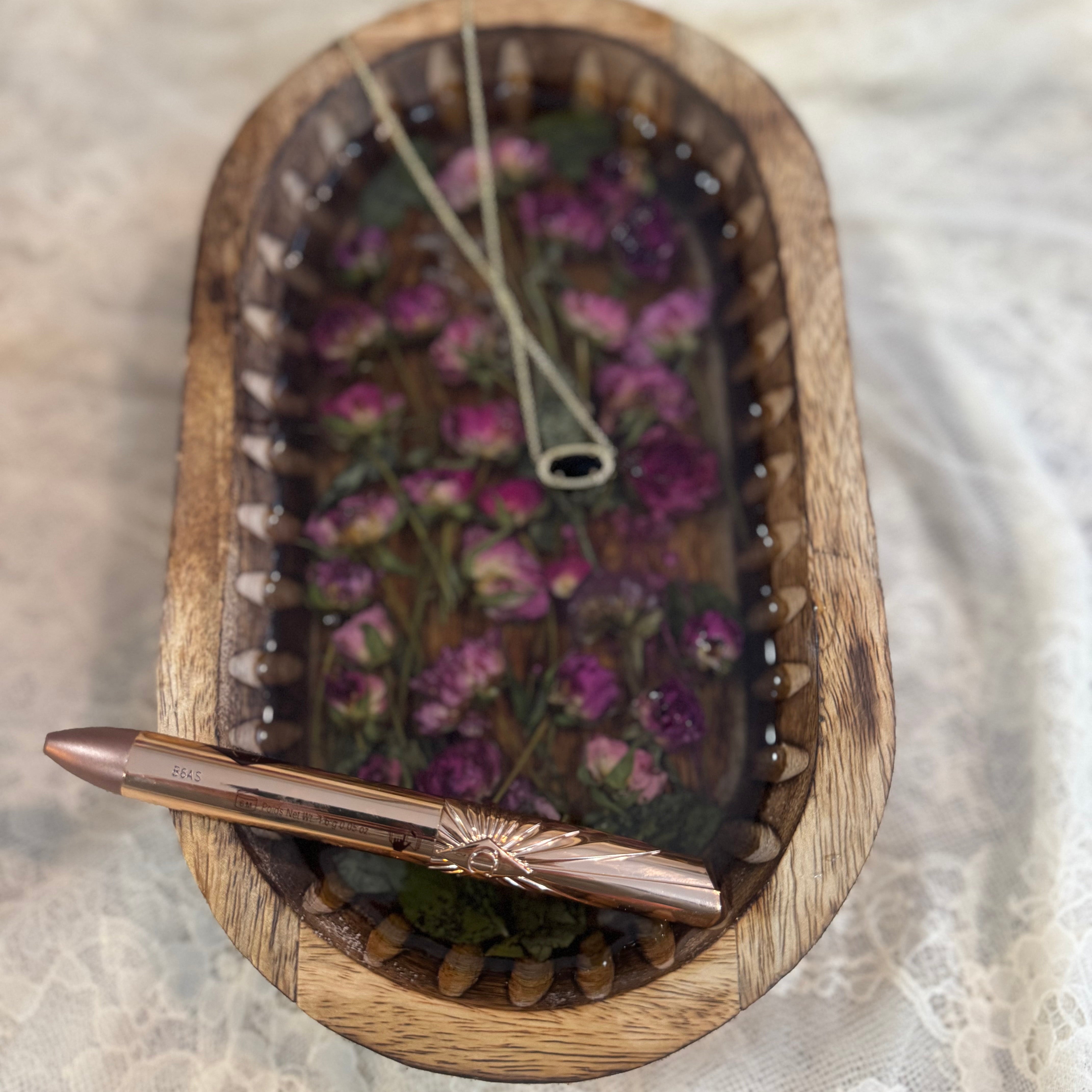 A close-up of an oval wooden tray filled with pressed purple flowers and greenery set in clear resin, on white lace fabric.