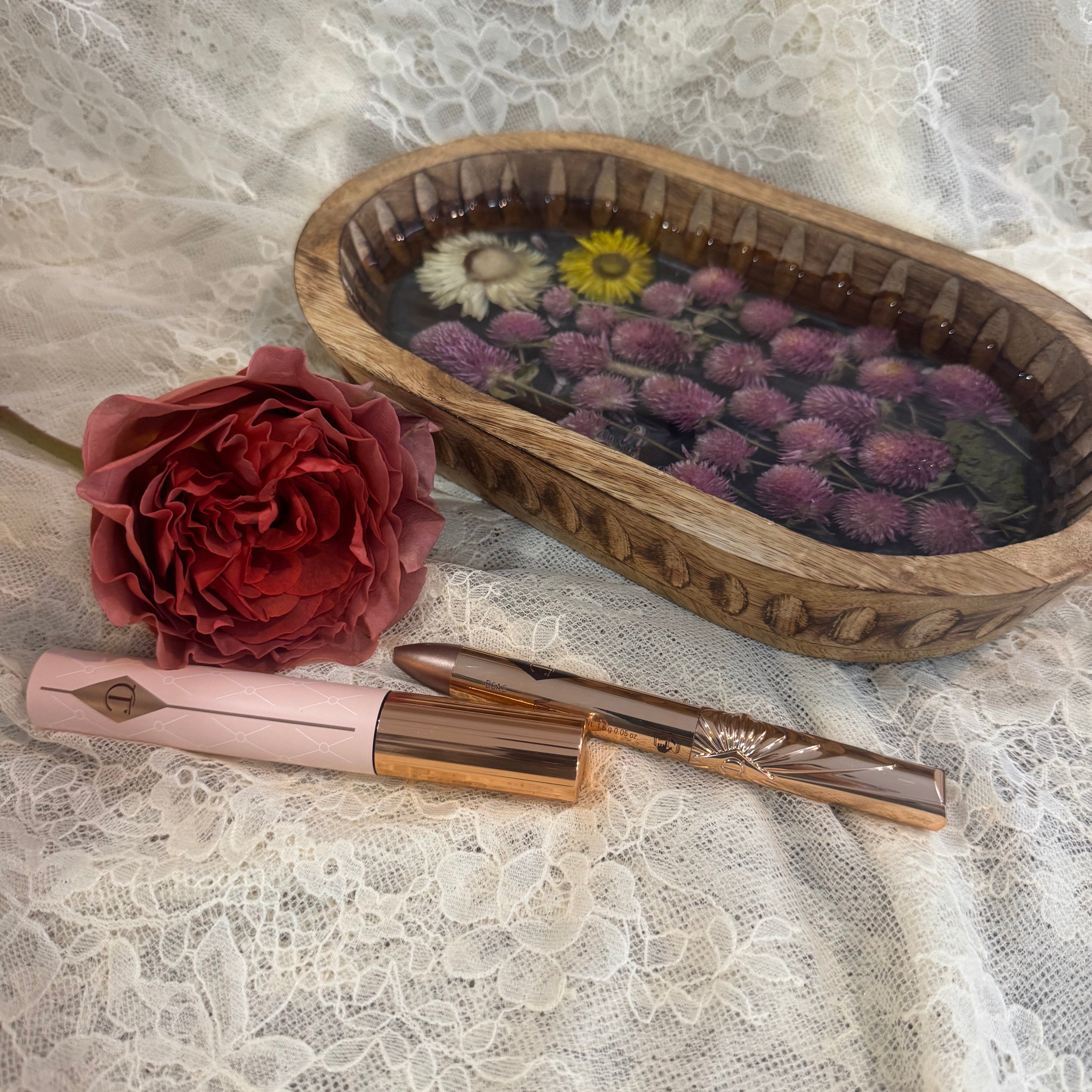 A pink rose and gold cosmetic pencils set  styled on white lace fabric next to a wooden tray containing dried flowers encased in clear resin.