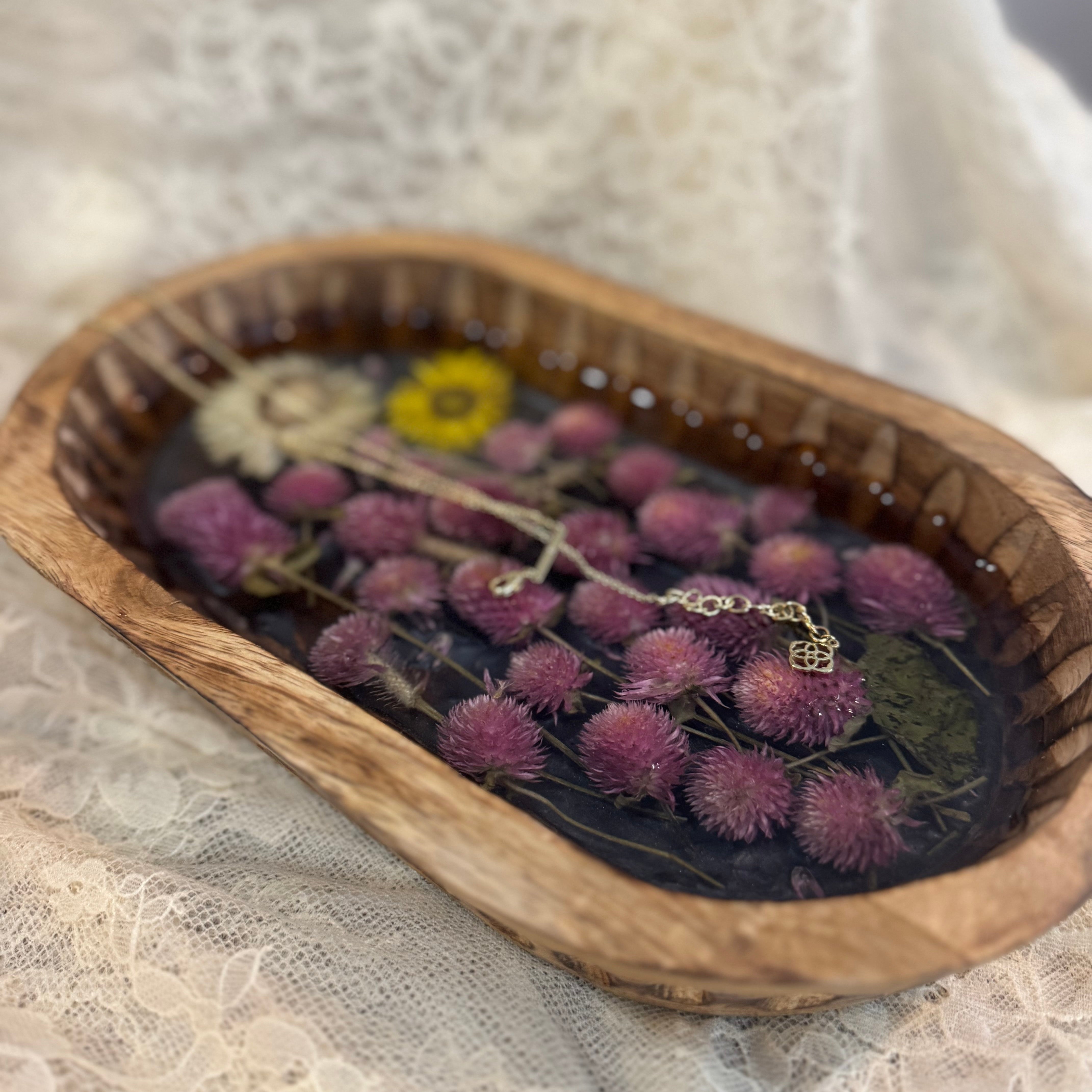 A small oval wooden tray filled with dried flowers and botanicals, including prominent pink-purple pom-pom flowers, resting on white lace fabric.