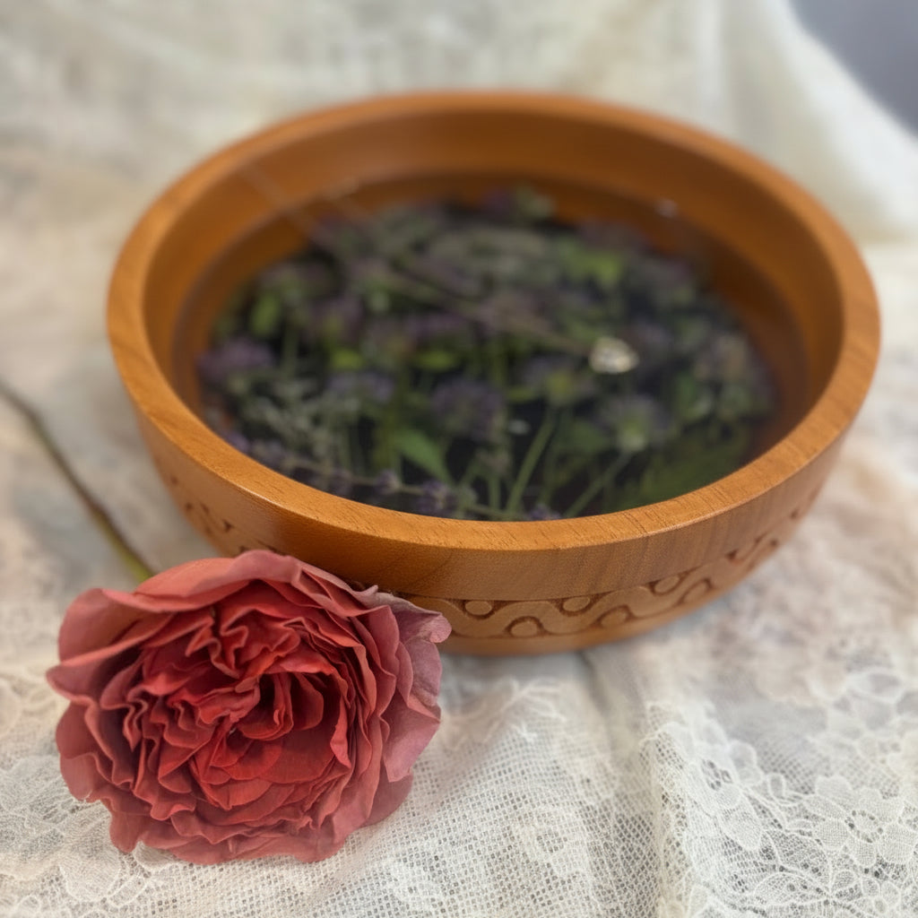 A carved wooden bowl filled purple clovers and floating green leaves, styled on a white lace cloth next to a single dried red rose.