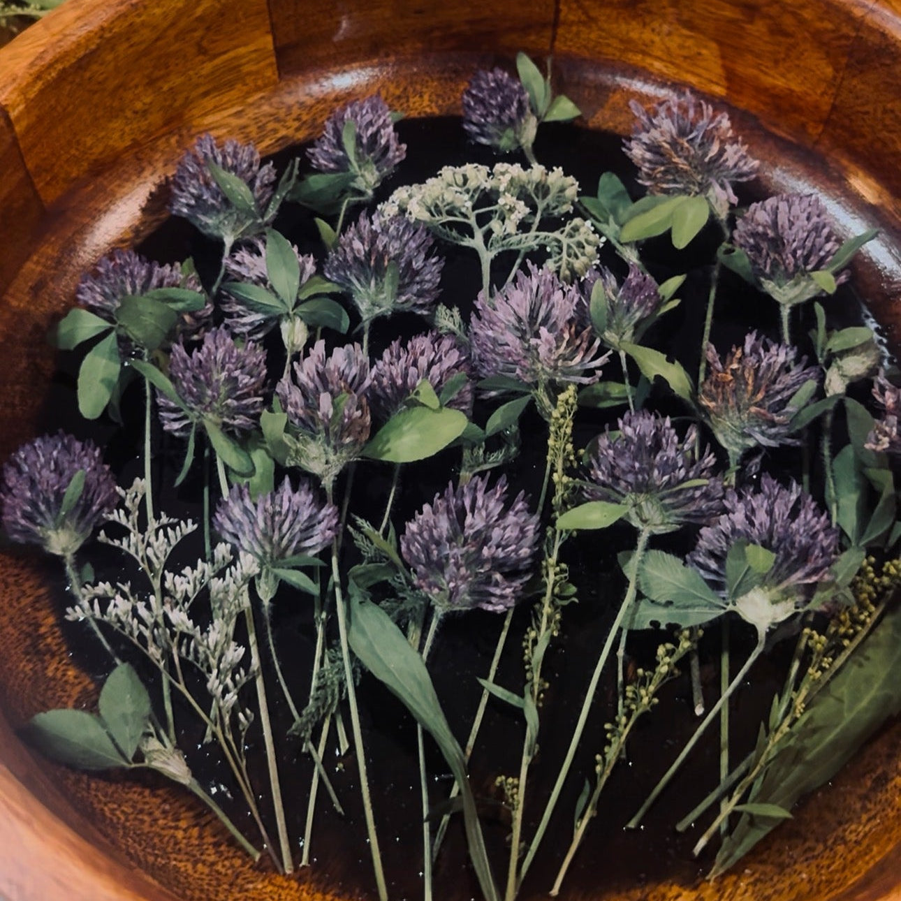 A wooden bowl filled with dried purple clover flowers and green leaves, with additional dried botanicals arranged in the background.