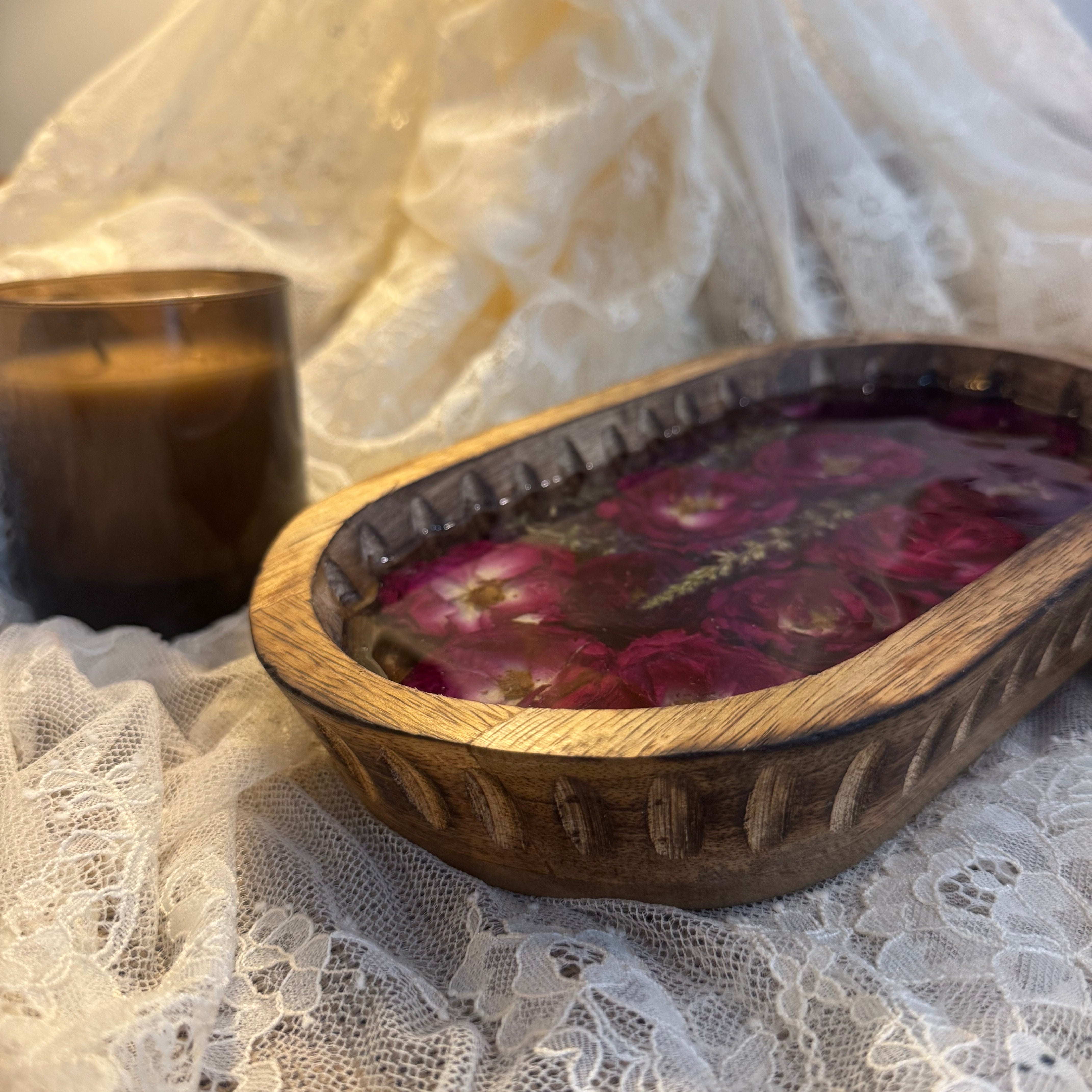 A rustic oval wooden bowl filled with dark red dried flowers and botanicals, styled on white lace fabric next to an amber glass candle jar.