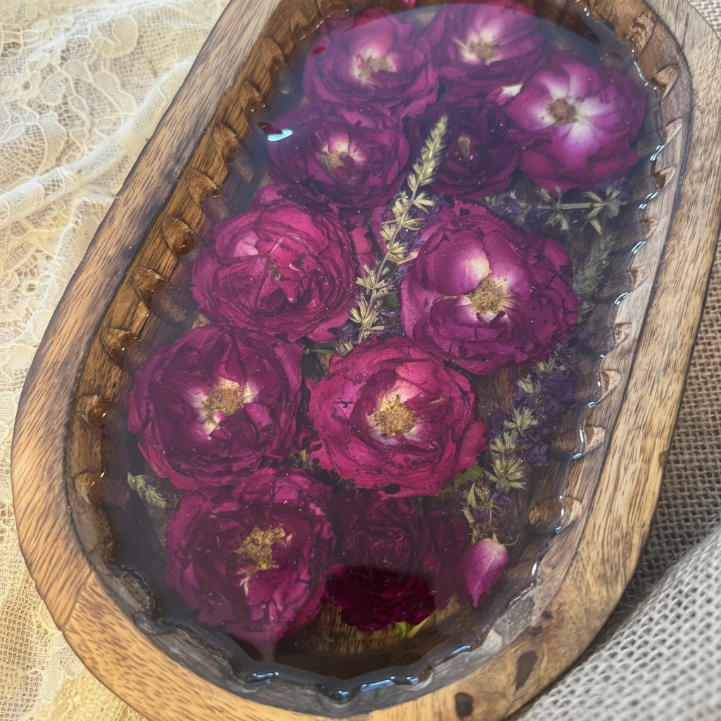 A decorative oval wooden tray filled with clear resin and preserved deep magenta-purple roses and small green botanicals, photographed on a lace cloth and burlap background.