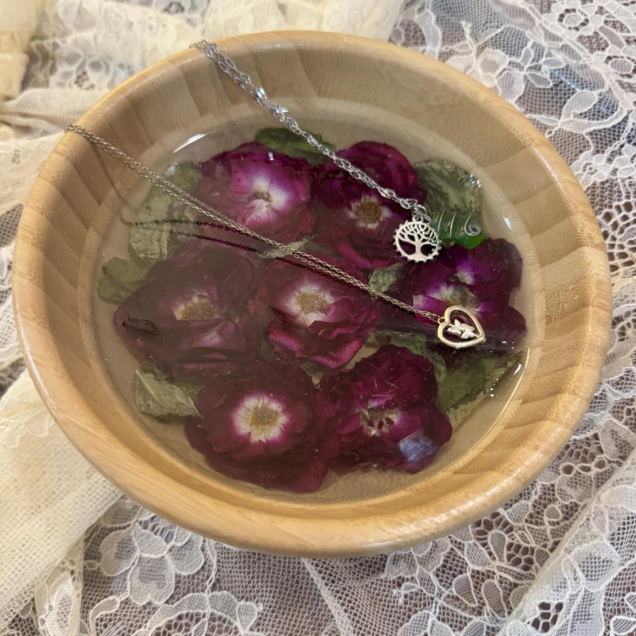Round bamboo trinket dish filled with clear resin and preserved burgundy roses, holding two delicate silver necklaces on white lace.