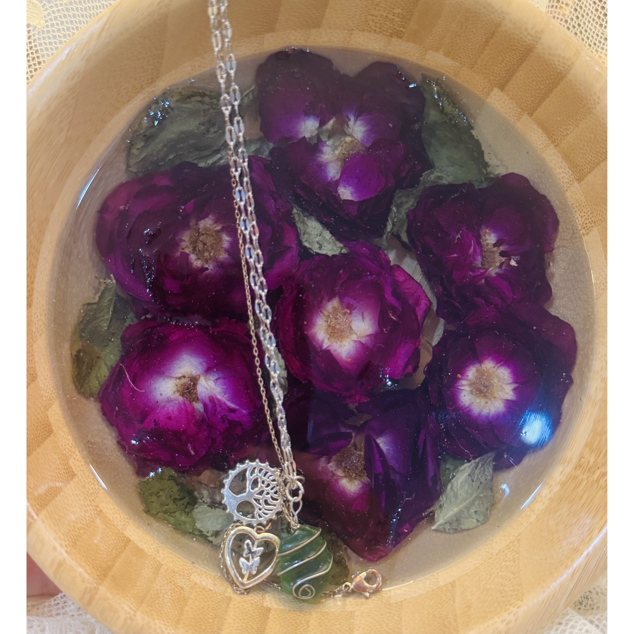 A round bamboo trinket dish filled with clear resin and preserved deep purple-burgundy roses and green leaves, holding a layered silver necklace with tree-of-life and heart charms and a wire-wrapped green stone.