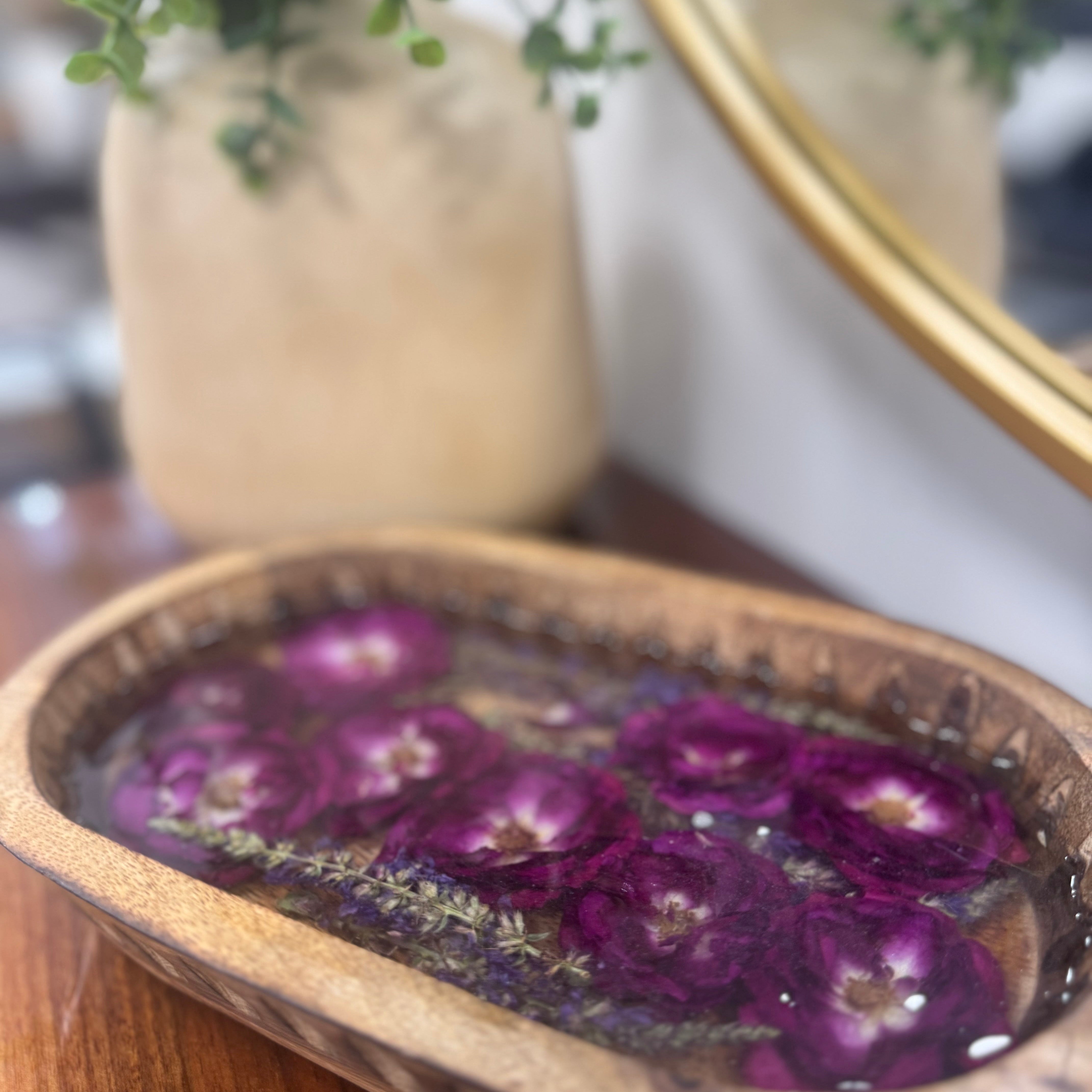 Carved oval mango wood dough bowl tray with deep magenta and purple preserved roses set in clear resin, on a wood surface with a gold-framed mirror behind