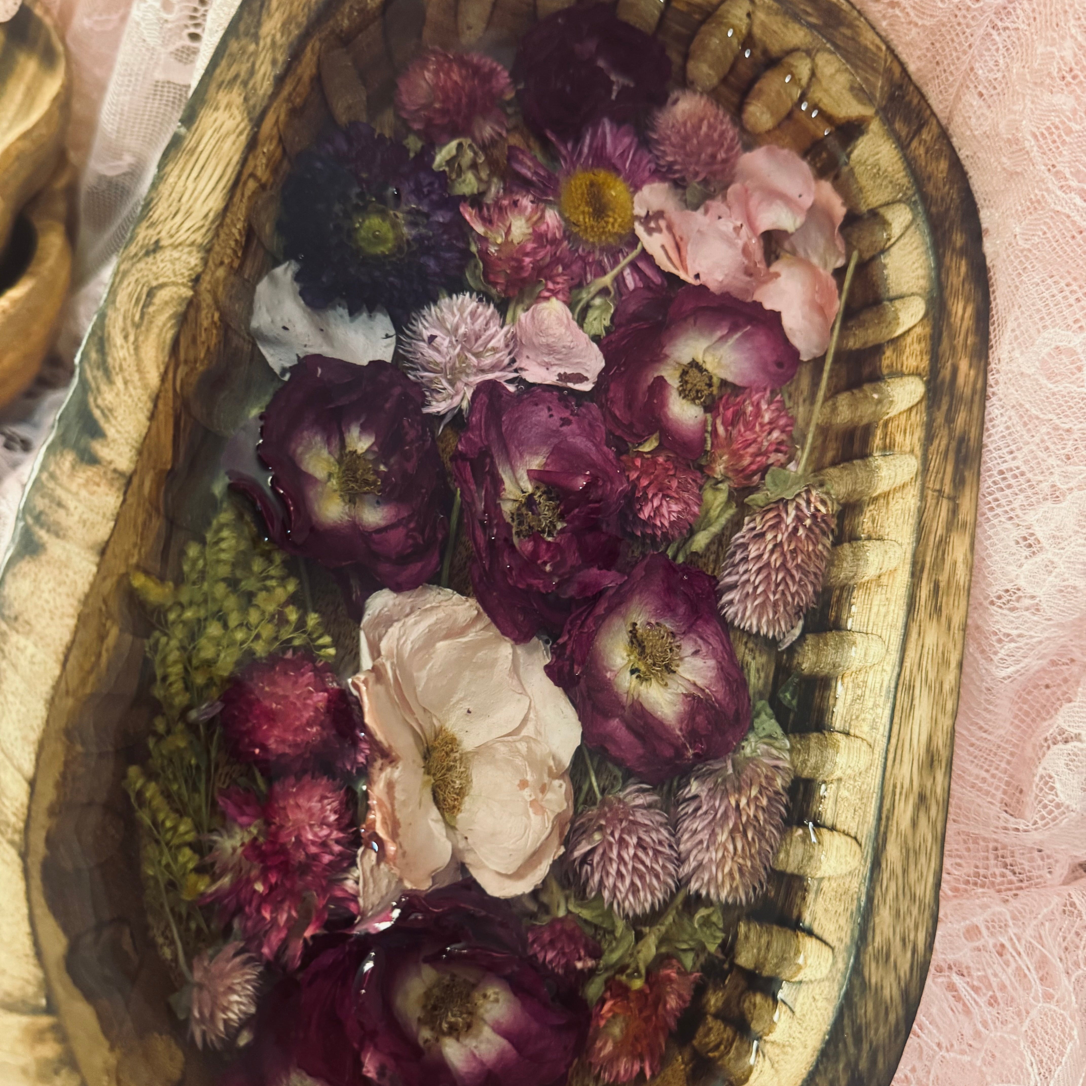 Close-up top-down view of a mango wood oval bowl with purple, pink, and cream preserved flowers and goldenrod set in glass-like resin