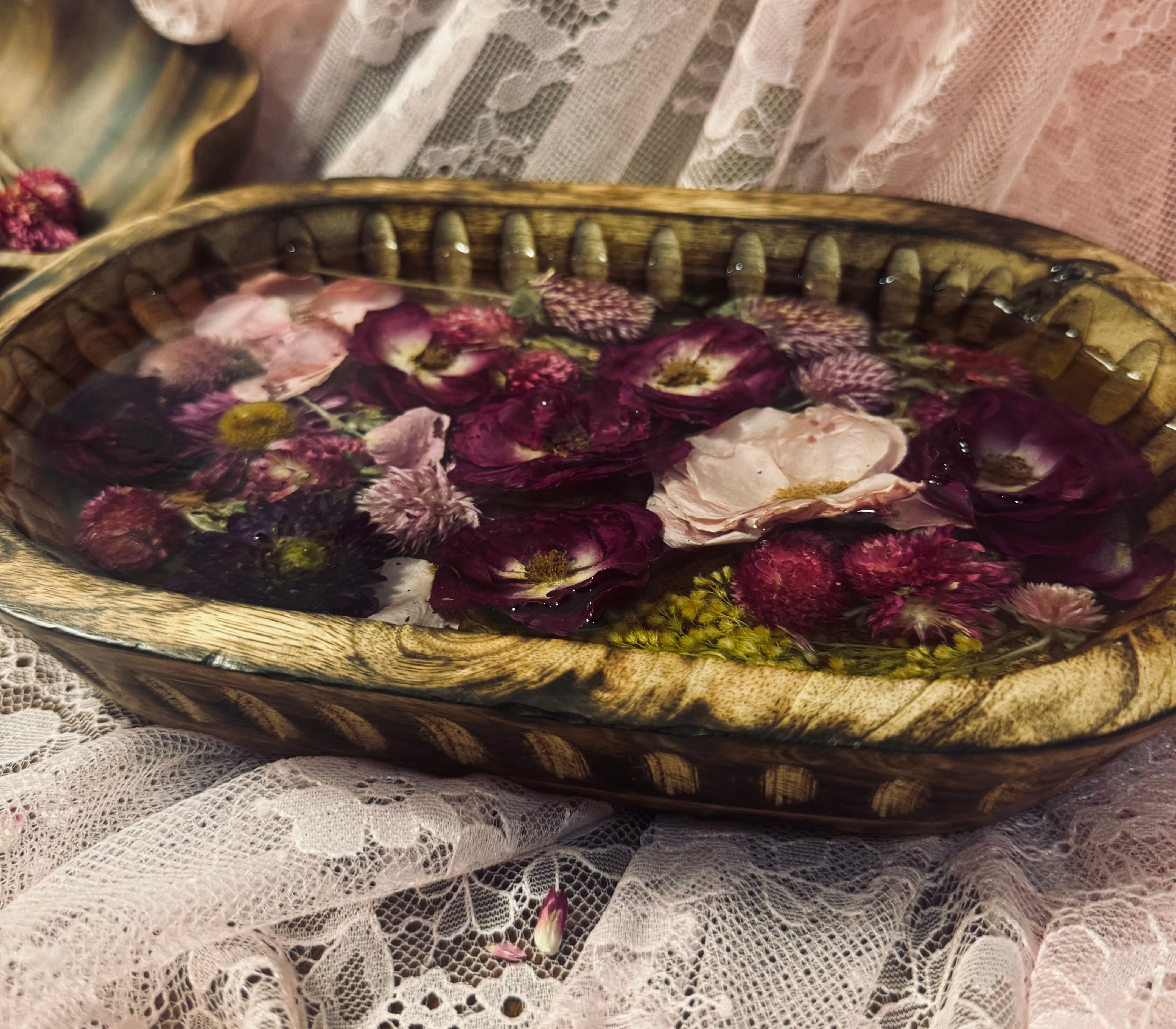 Oval wood bowl filled with layered pink, burgundy, and lavender dried flowers sealed in glass-like resin, styled on pink lace