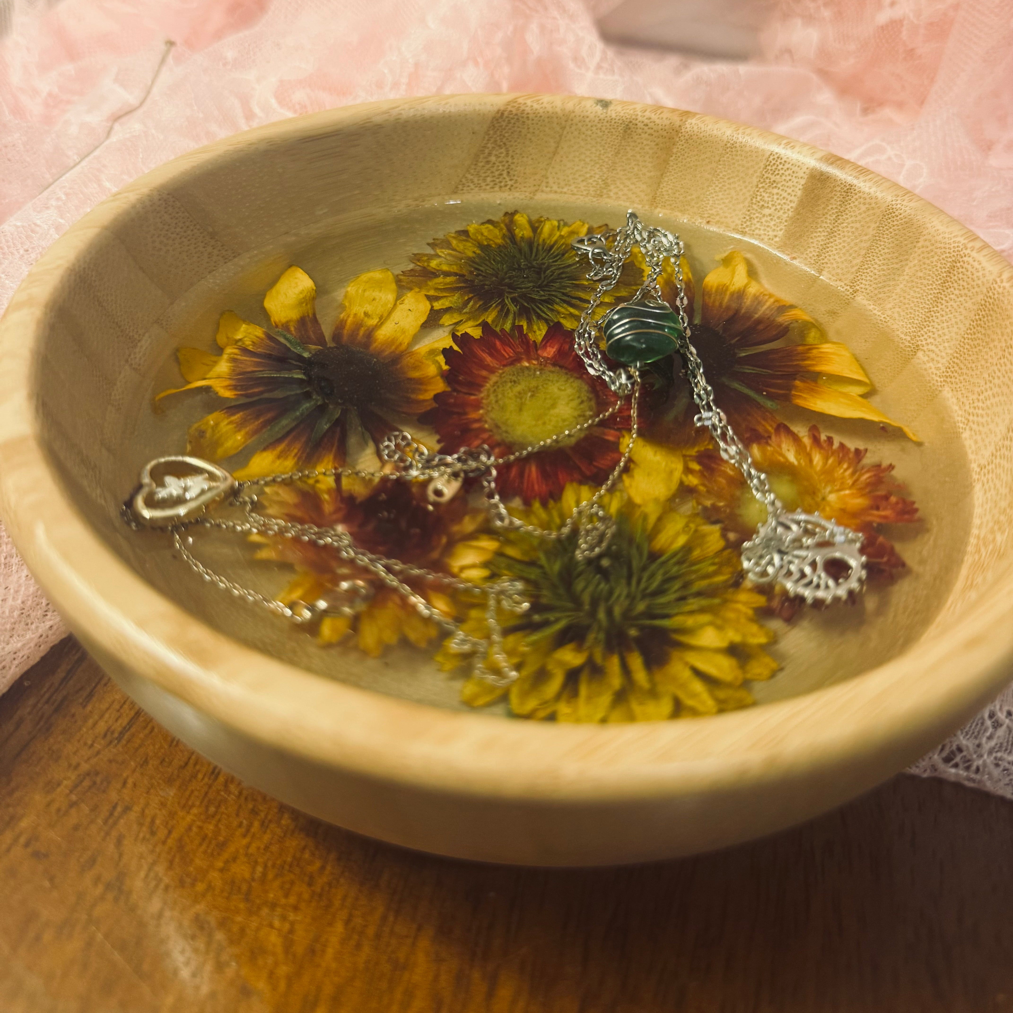 Close-up top-down view of a round bamboo bowl with golden and burgundy strawflowers preserved in glass-like resin, styled with silver necklaces on a wood tabletop