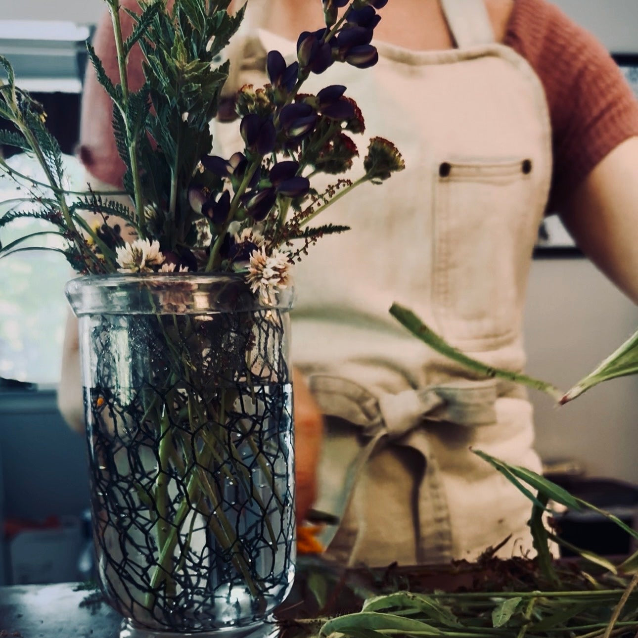 A florist in a denim apron arranging fresh purple lupine and white clover into a glass jar of water, with loose stems scattered on the worktable