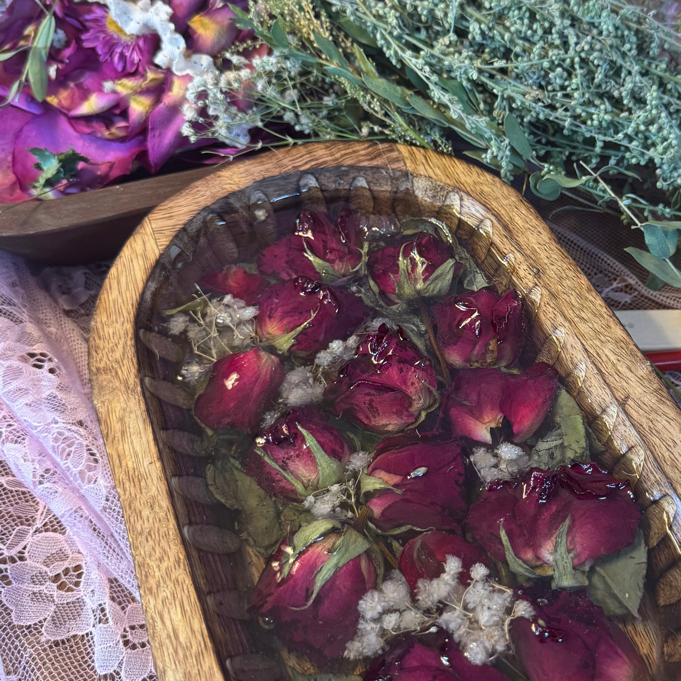 A wooden oval tray filled with dried red rose buds and green leaves, styled on pink lace fabric with dried flowers in the background.