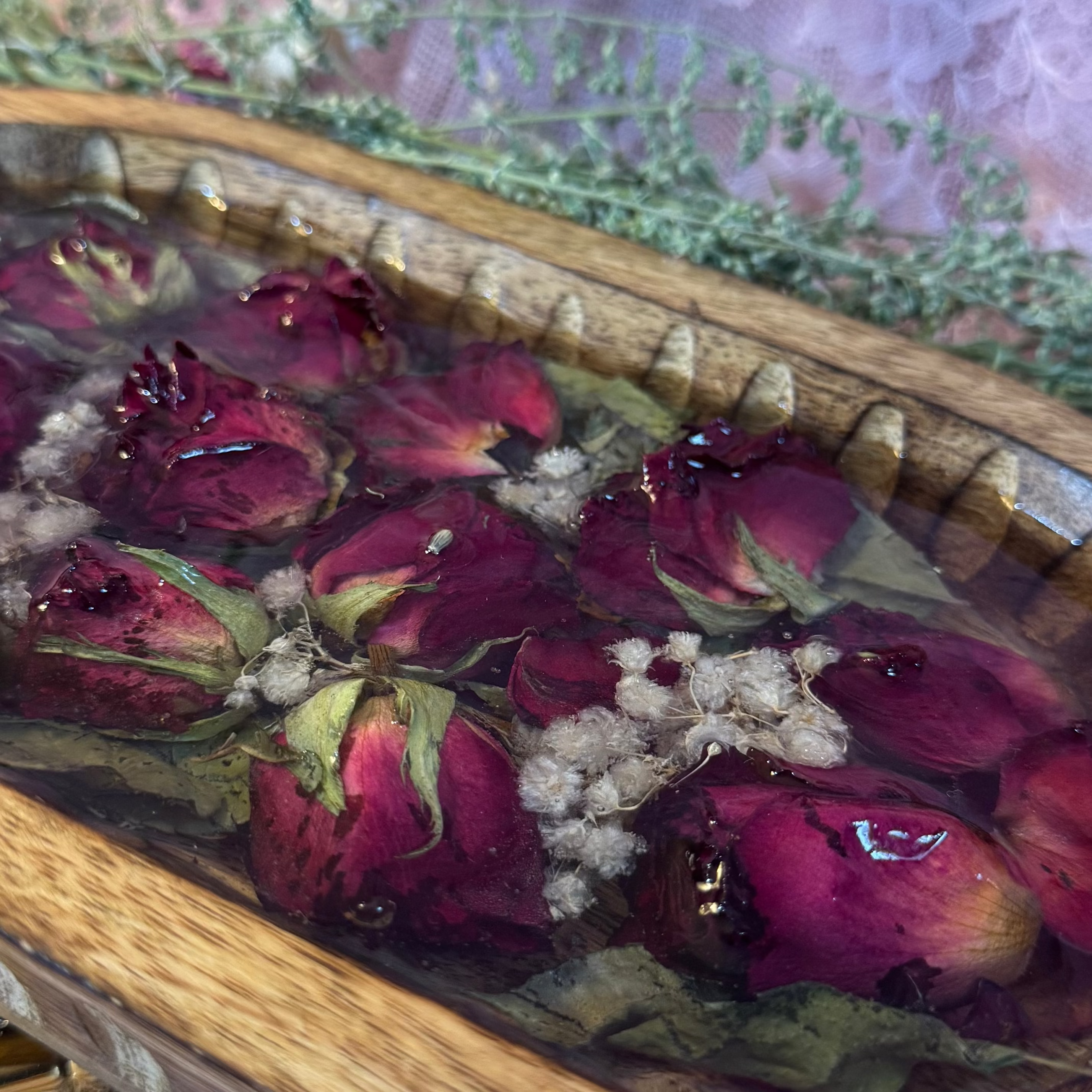A wooden oval bowl filled with glass like resin with floating dried rose buds, green leaves, and small white flowers, styled on lace fabric with a pink lace backdrop.