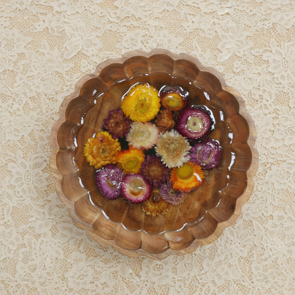A close-up of a scalloped wooden bowl with dried wildflowers preserved in resin, styled on a light beige textured surface.