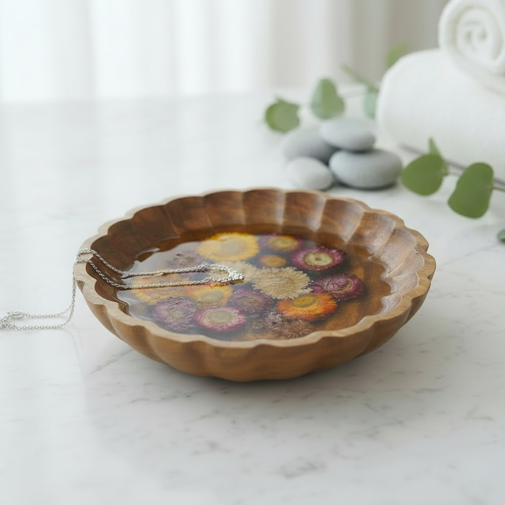 A scalloped wooden bowl filled with dried wildflowers set in clear resin, styled next to makeup and beauty products on a light beige surface.